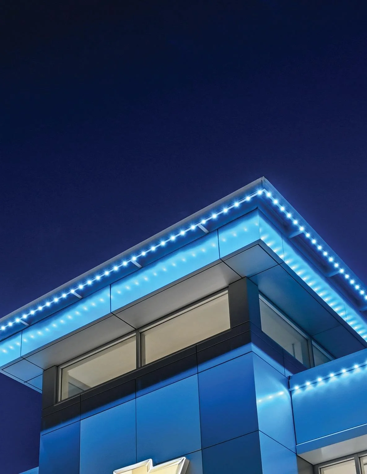 Permanent lighting system, automative, nighttime view of a modern building with blue exterior panels, large glass windows, and blue LED strip lighting along the roofline.