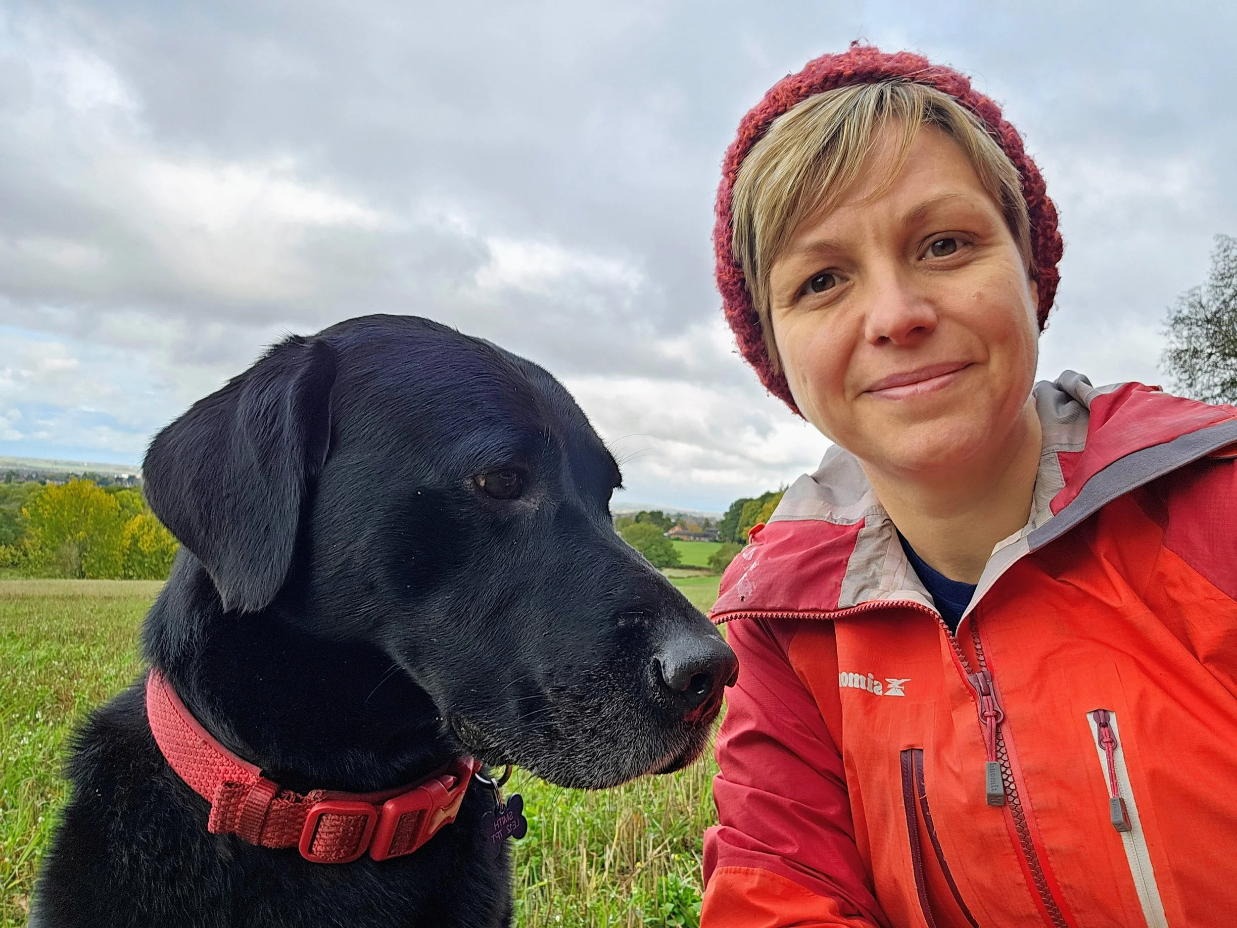 A black Labrador sitting next to Kat from Kat’s Dogs during a countryside walk in Loughborough, both looking relaxed and content