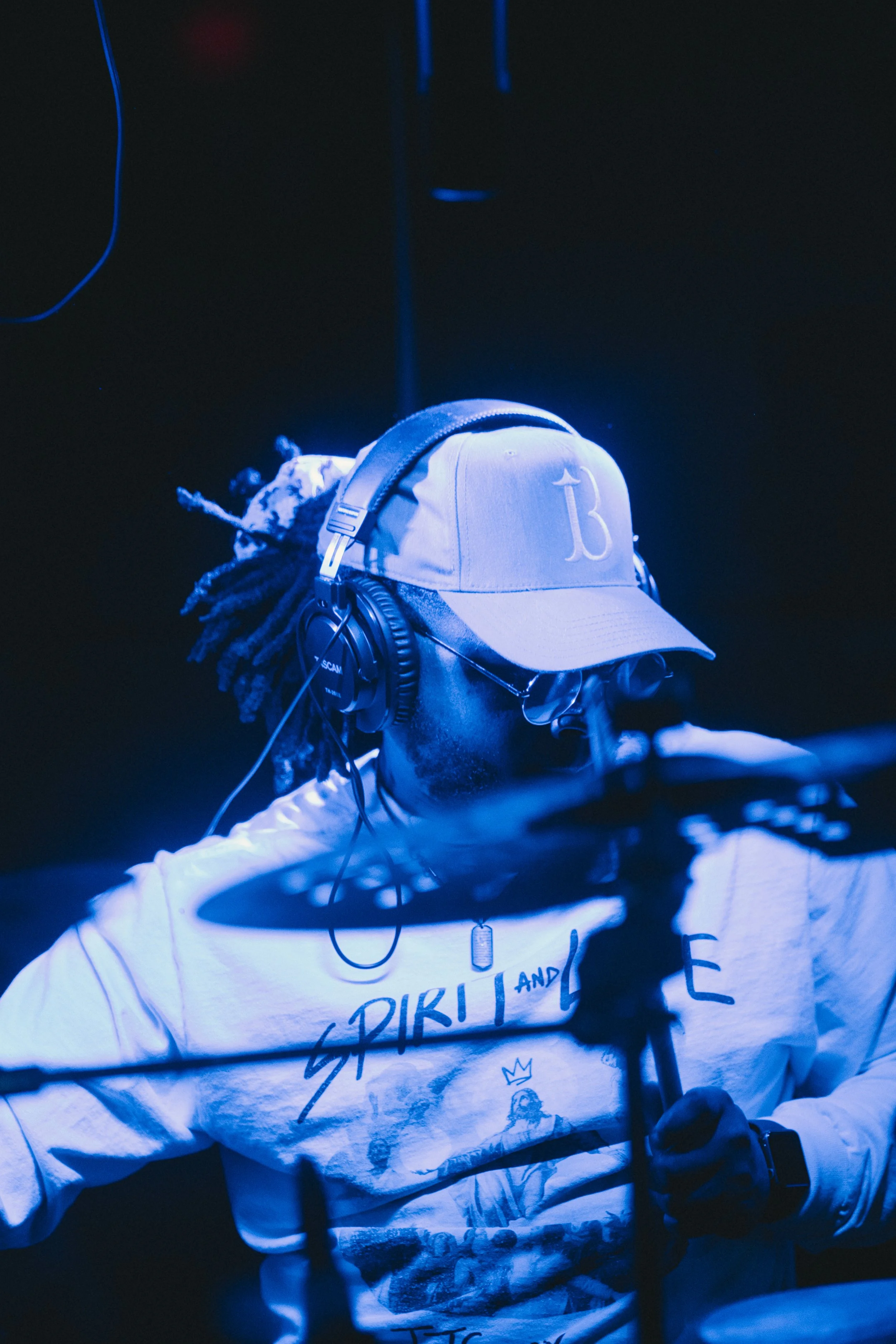 A DJ with dreadlocks, wearing a baseball cap, sunglasses, and headphones, performing behind a drum kit in a dimly lit setting.