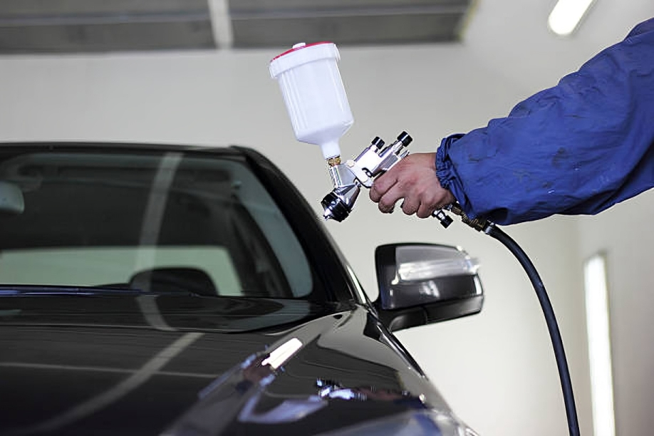 A person wearing a blue jacket is using a paint spray to repair a car window in a garage.