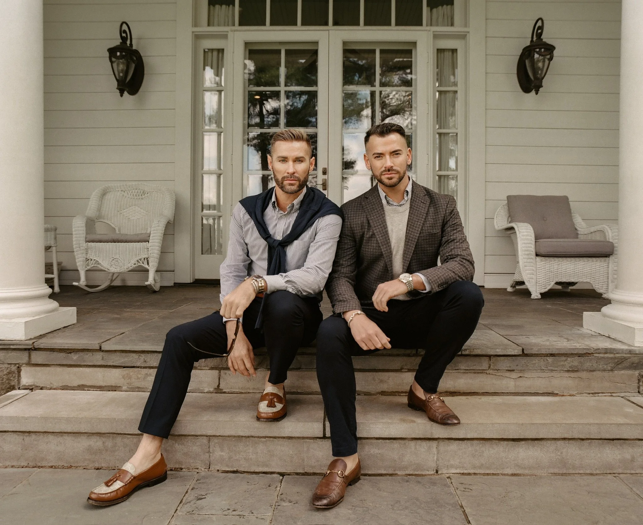 Two men sitting on the steps of a porch in front of a white house with double glass doors and two black outdoor lamps. They are dressed in business casual attire.
