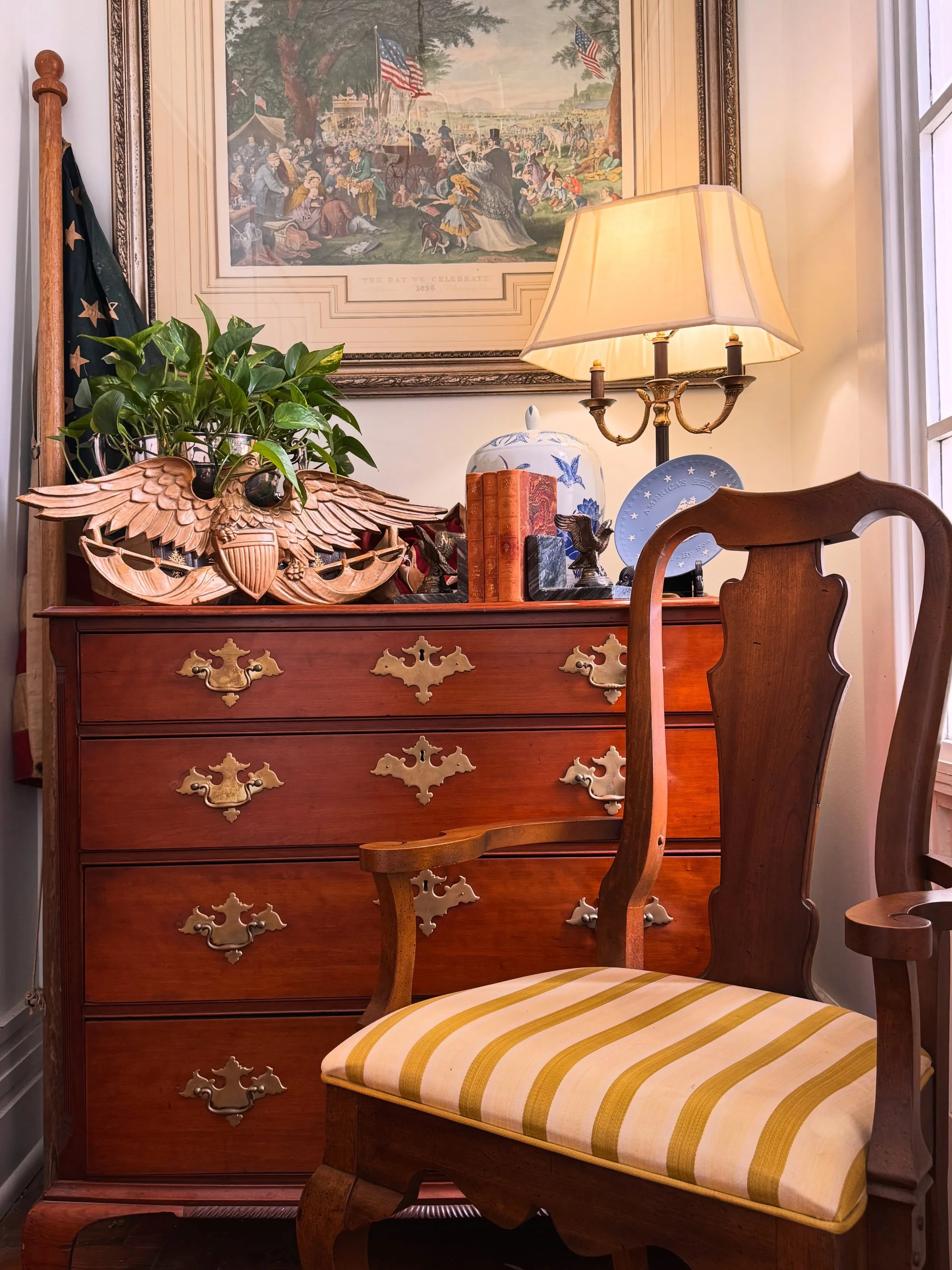 A wooden dresser with ornate brass handles, holding a potted plant, two eagle figurines, a decorative pitcher, and books. Behind it, a framed historical print hangs on the wall, illuminated by a table lamp with a beige shade. There is a wooden chair with a yellow-striped upholstery in the foreground.