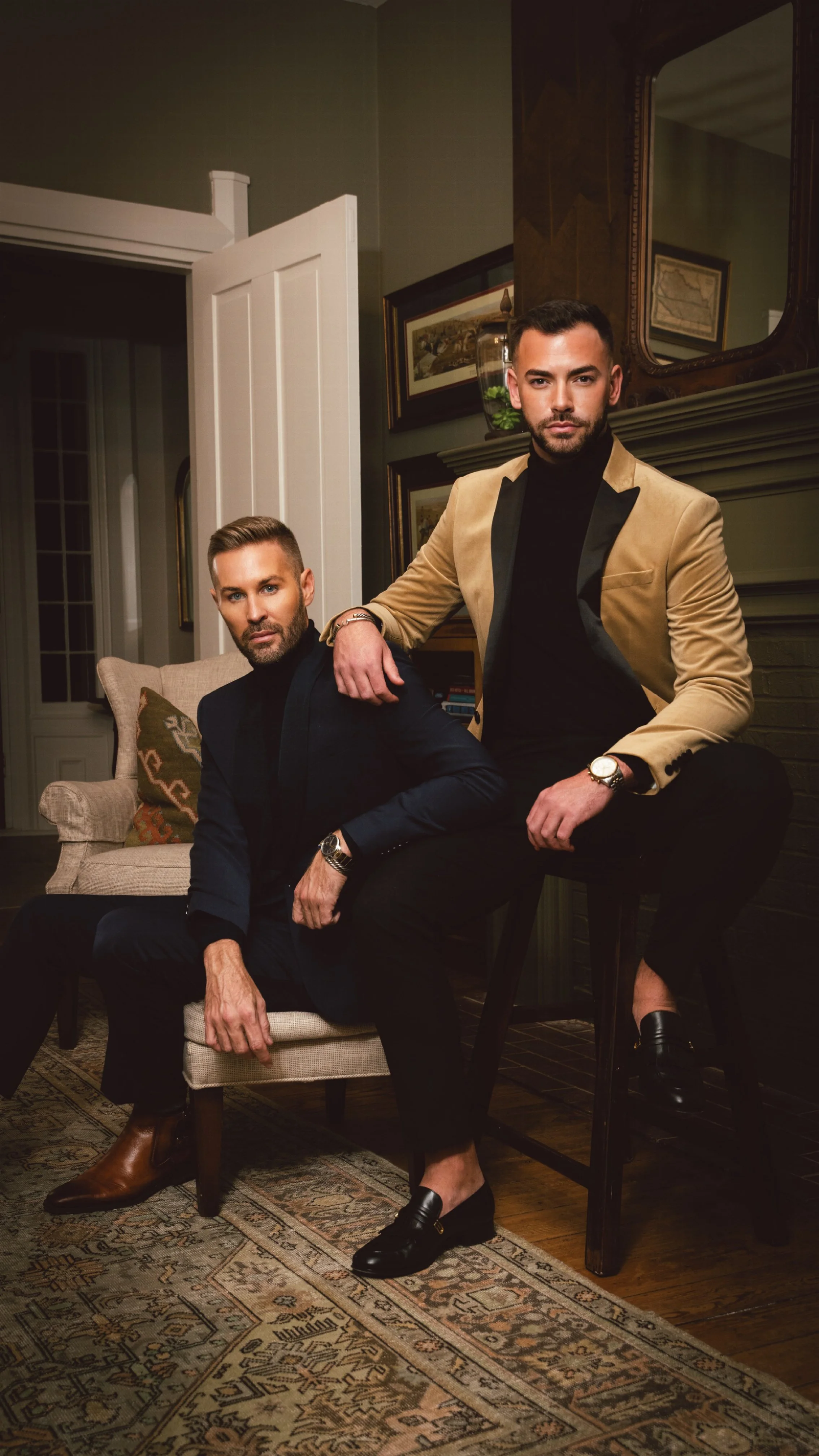 Two men posing in a stylishly decorated living room, sitting and standing on furniture with artwork and a mirror in the background.