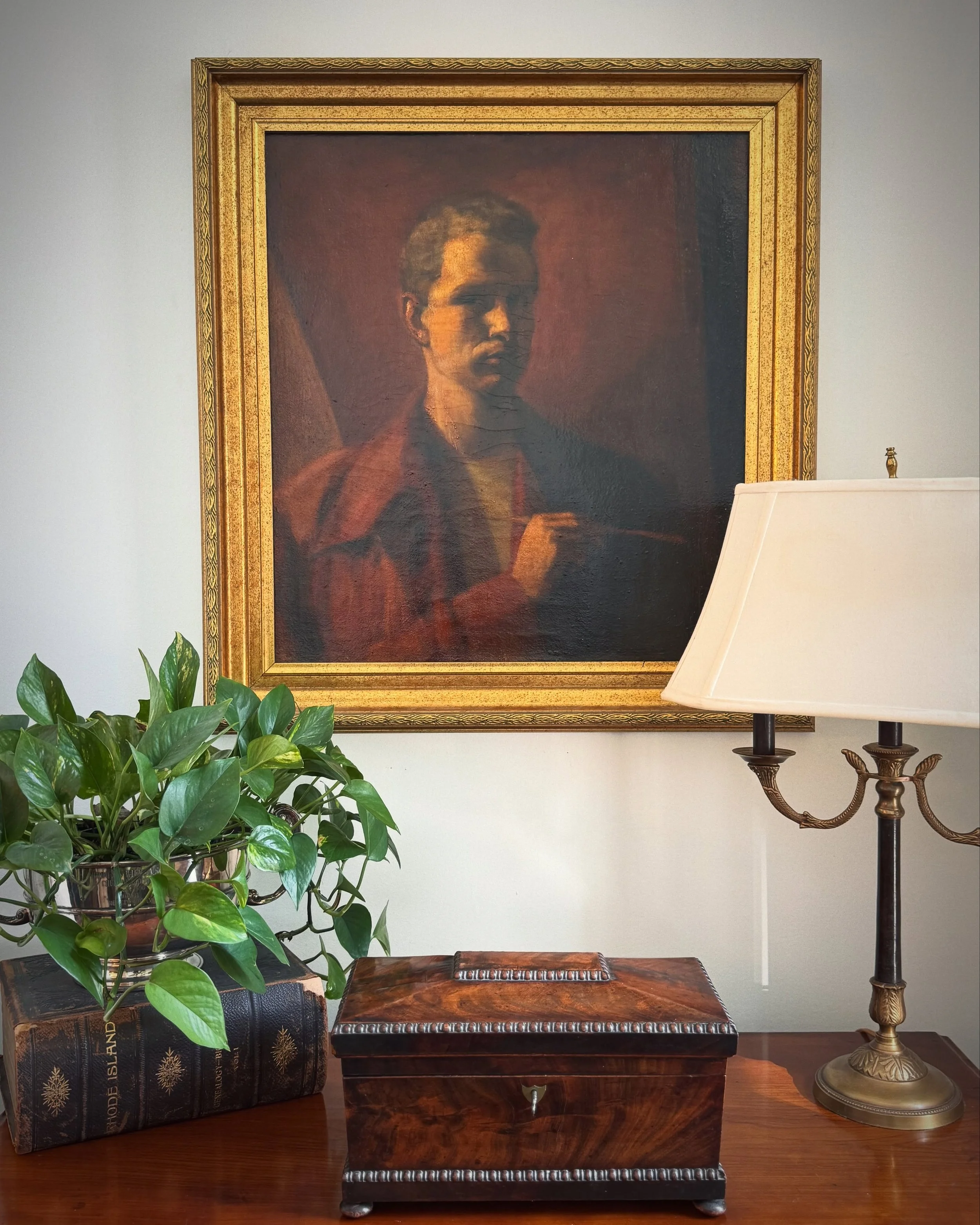 A decorative living room corner featuring a framed portrait painting of a woman, a green potted plant, a wooden chest, and a brass lamp with a white shade on a side table.