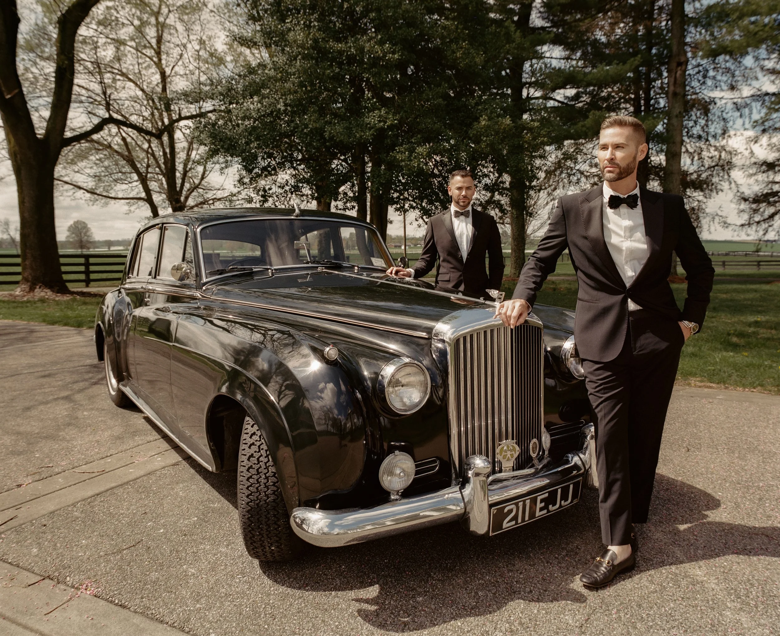 Two men in black tuxedos and bow ties stand beside a vintage black car with a woman standing nearby, in an outdoor setting with trees and open fields in the background.