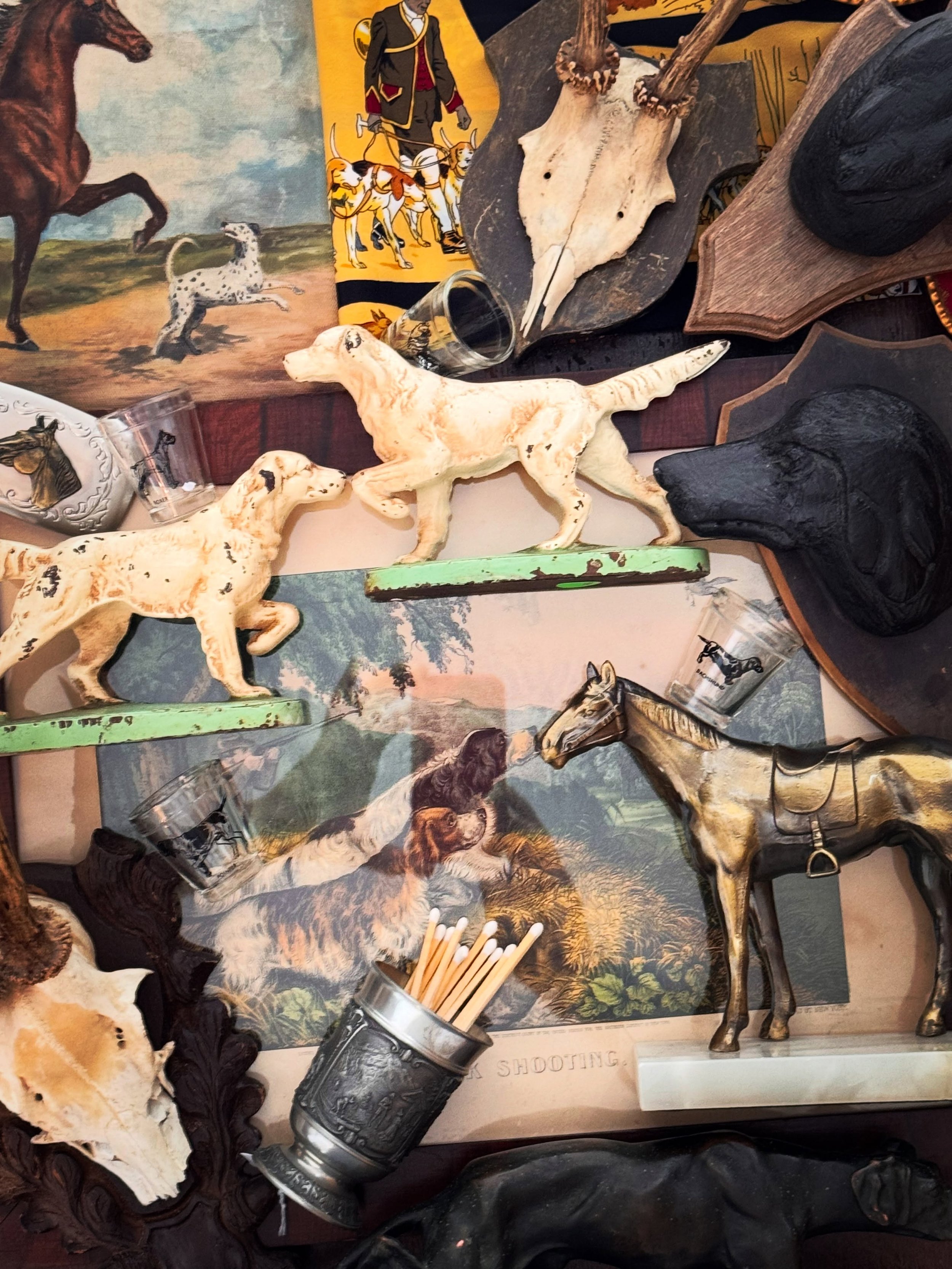 Collection of hunting-themed items, including carved wooden animal figures such as a dog and a horse, animal skulls, hunting-themed artwork, and hunting accessories like shot glasses and arrow-shaped pens.