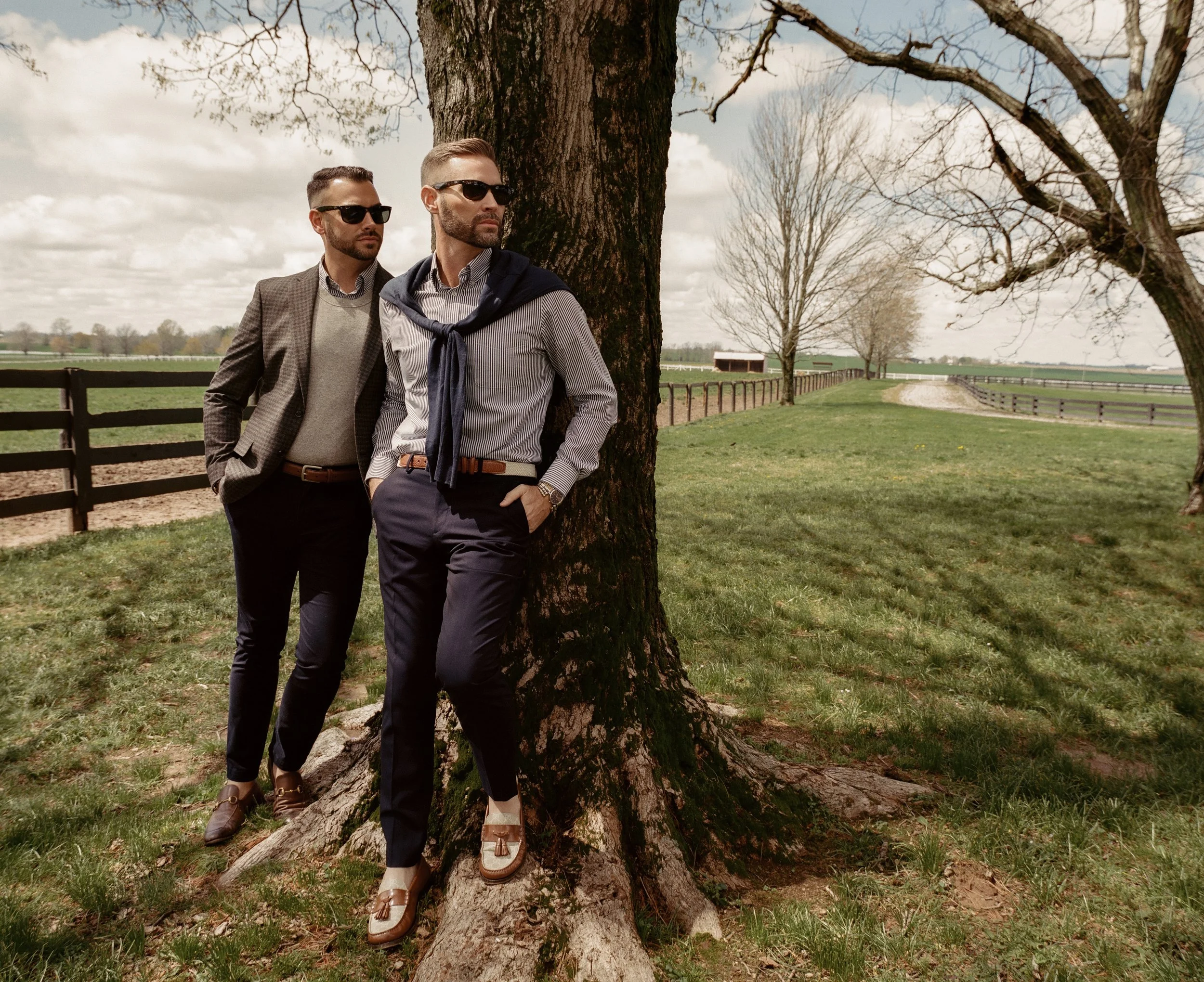 Two men with sunglasses and stylish clothing leaning against a tree in a rural landscape with green fields, a fence, and budding trees, on a partly cloudy day.