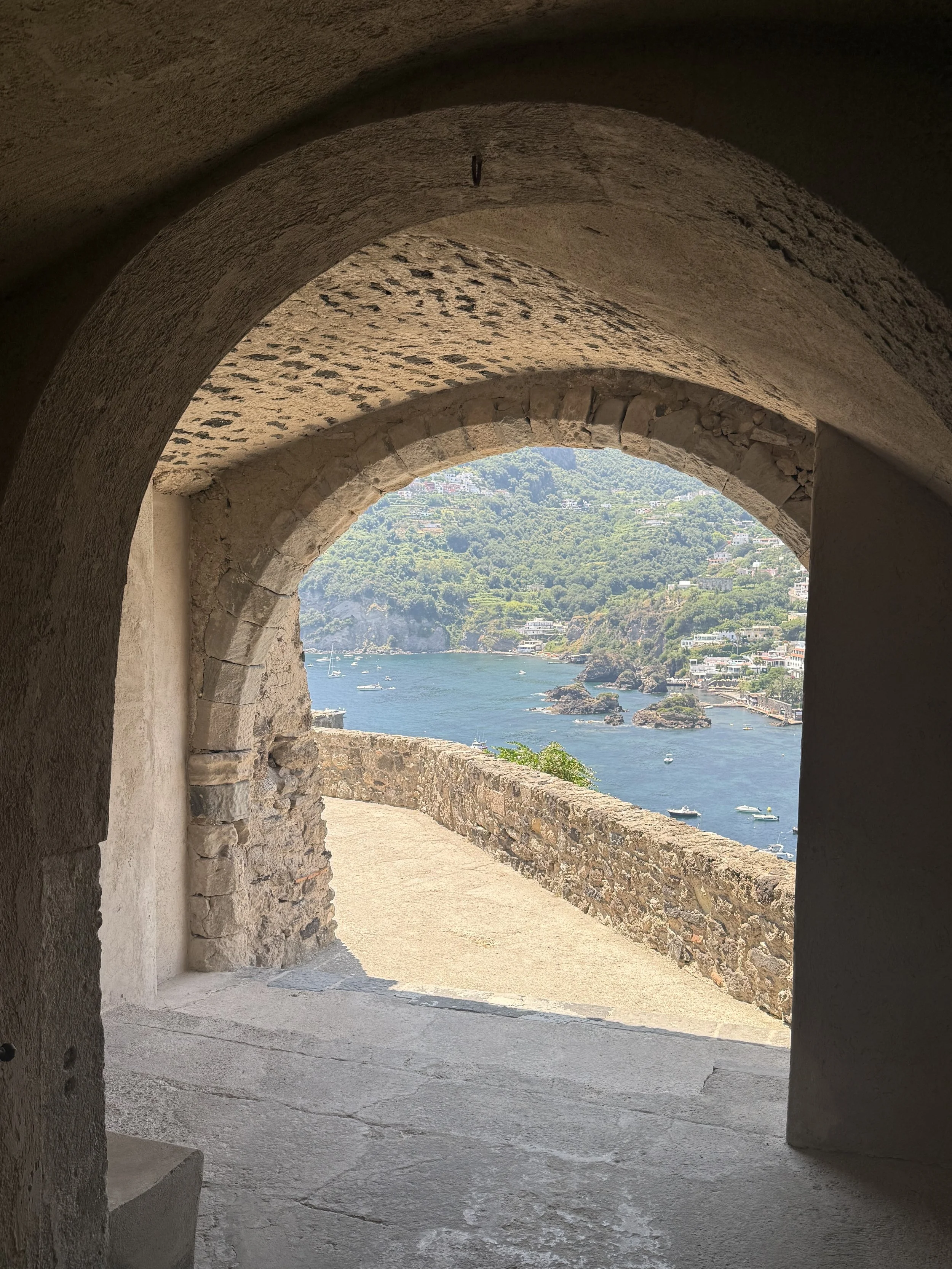 View of a coastal landscape through an arched stone window, with boats on the water and green hills in the background.