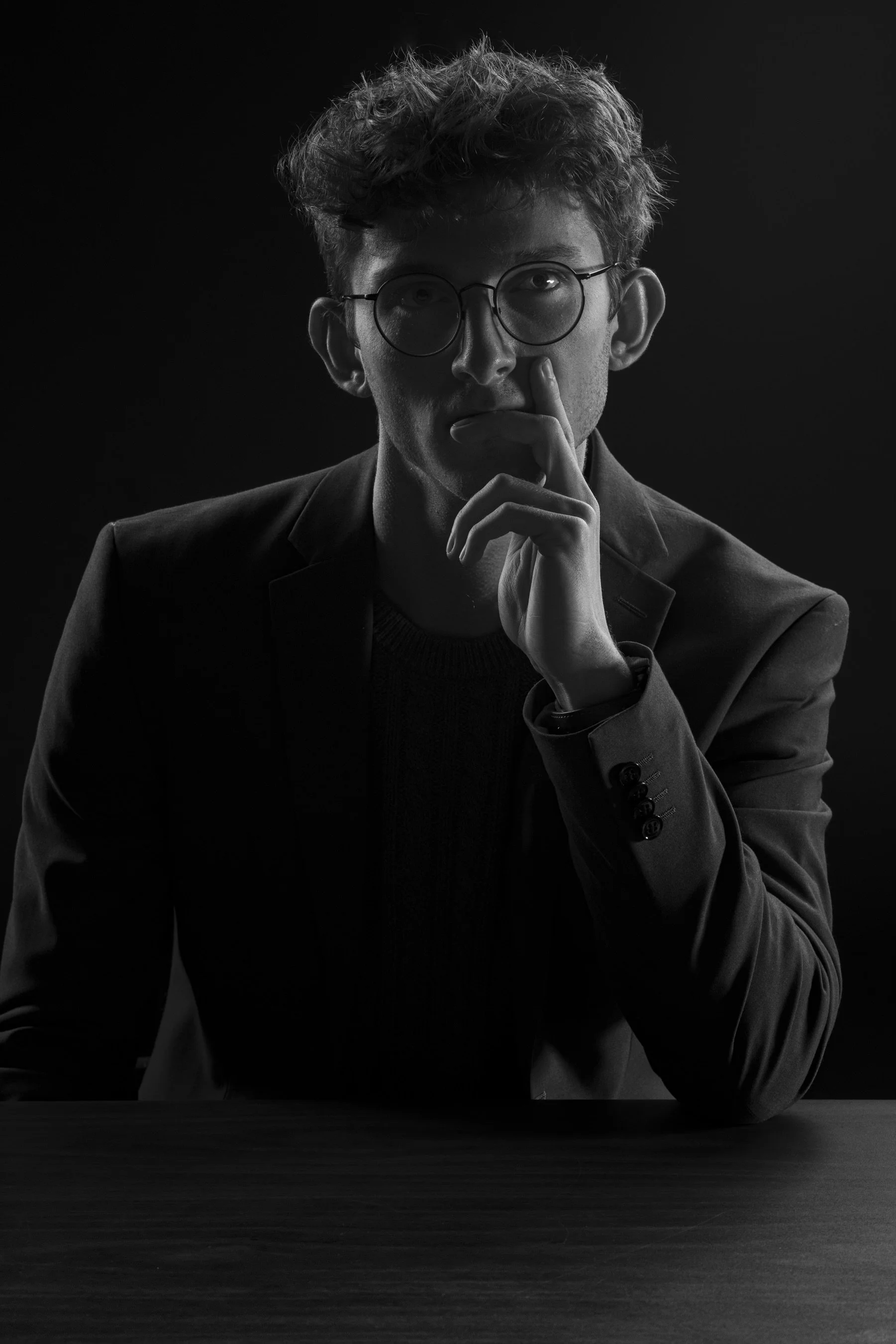 Black and white portrait of a young man with curly hair and glasses, resting his chin on his hand in a thoughtful pose, sitting at a table.