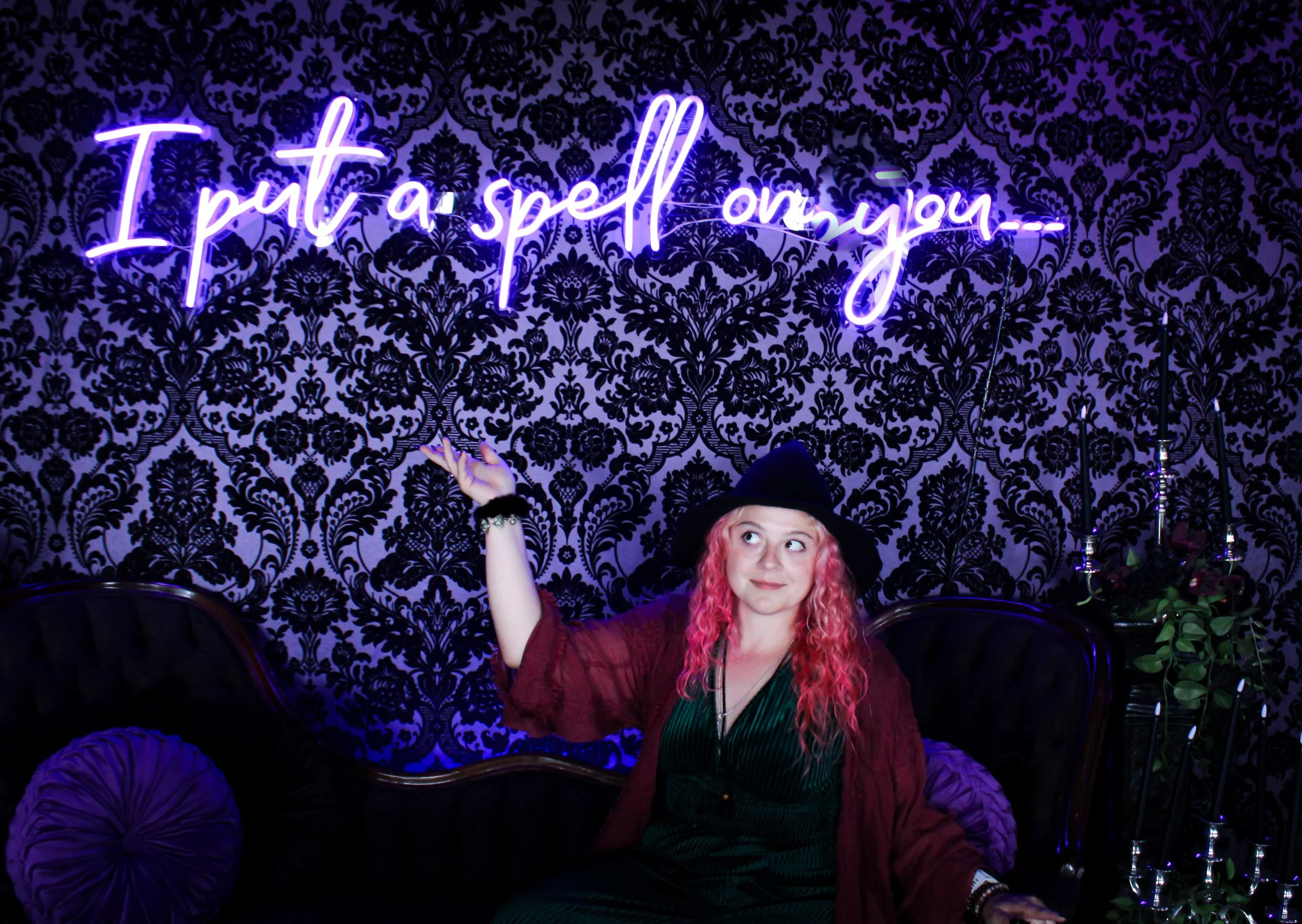 A woman with pink curly hair wearing a black witch hat, green dress, and maroon shawl sitting on a black velvet sofa, pointing at a neon sign that says "I put a spell on you" in purple, hanging on a black floral damask patterned wall. In Salem.