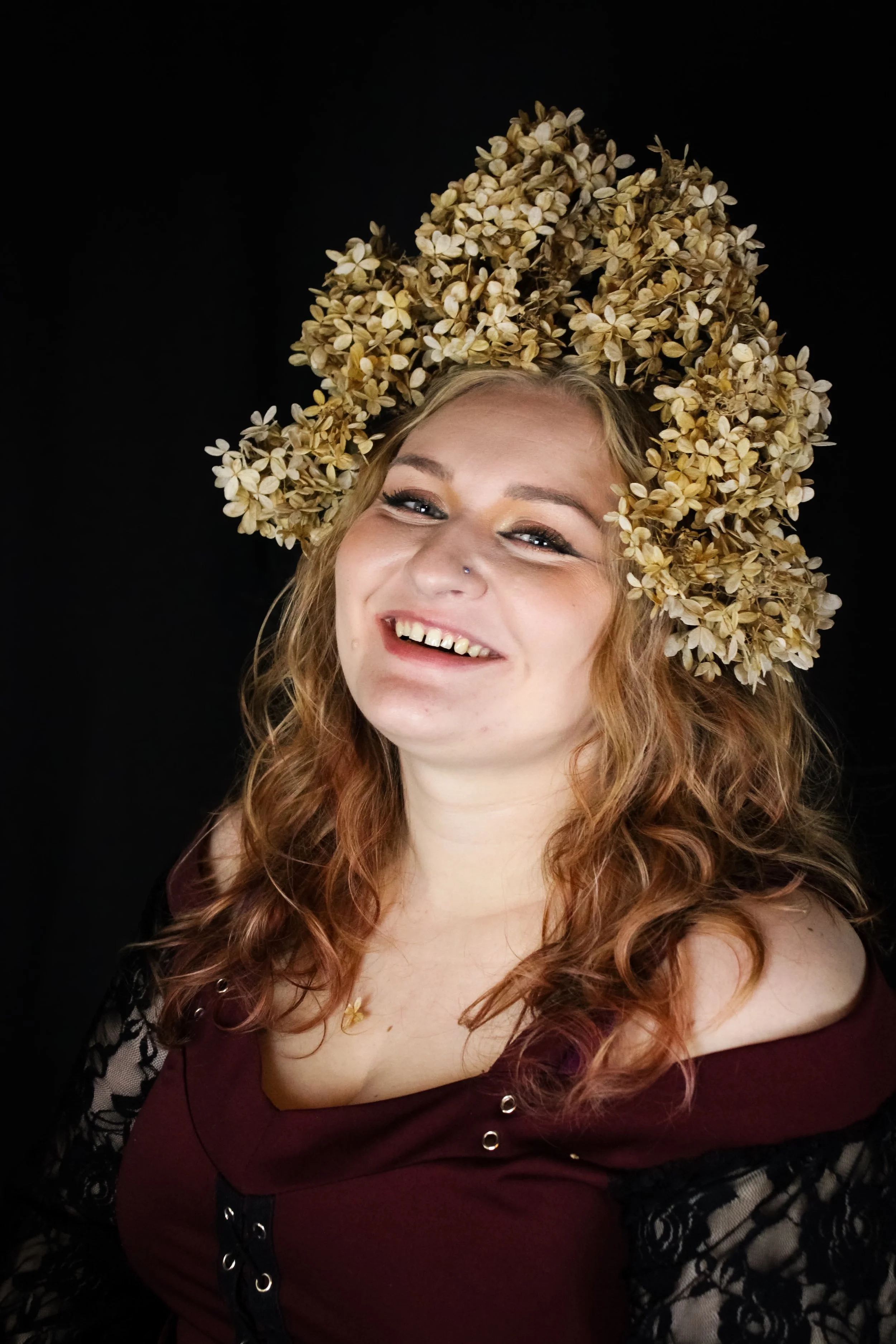 A woman with curly red hair wearing a floral headdress made of dried flowers, smiling against a black background.