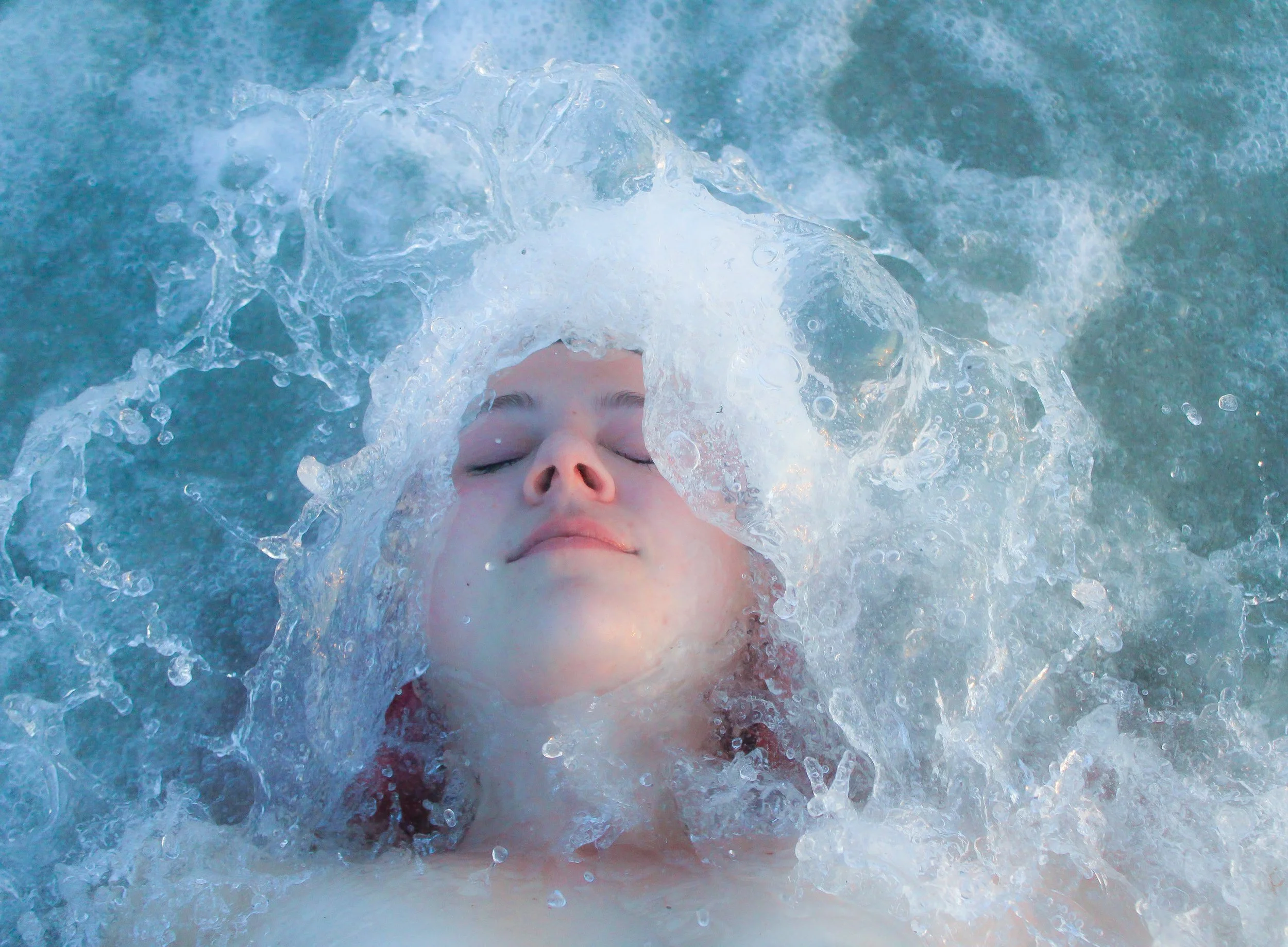 A person submerged in water with their eyes closed, surrounded by splashing water. Waves of the ocean crashing onto woman as she soaks it in.
