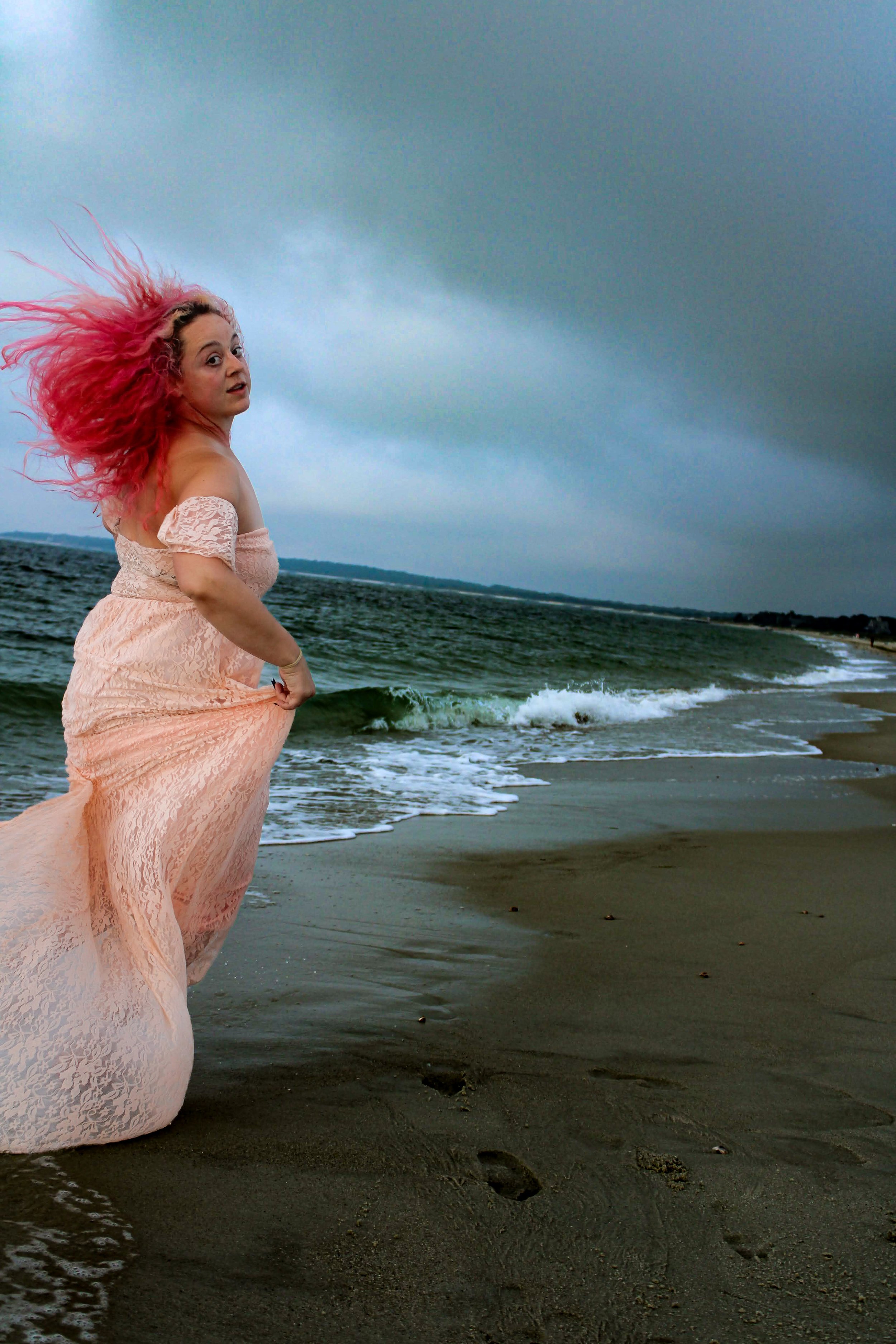 A woman with bright pink hair running on a sandy beach, wearing a light pink lace dress, with dark, cloudy sky and ocean waves in the background.