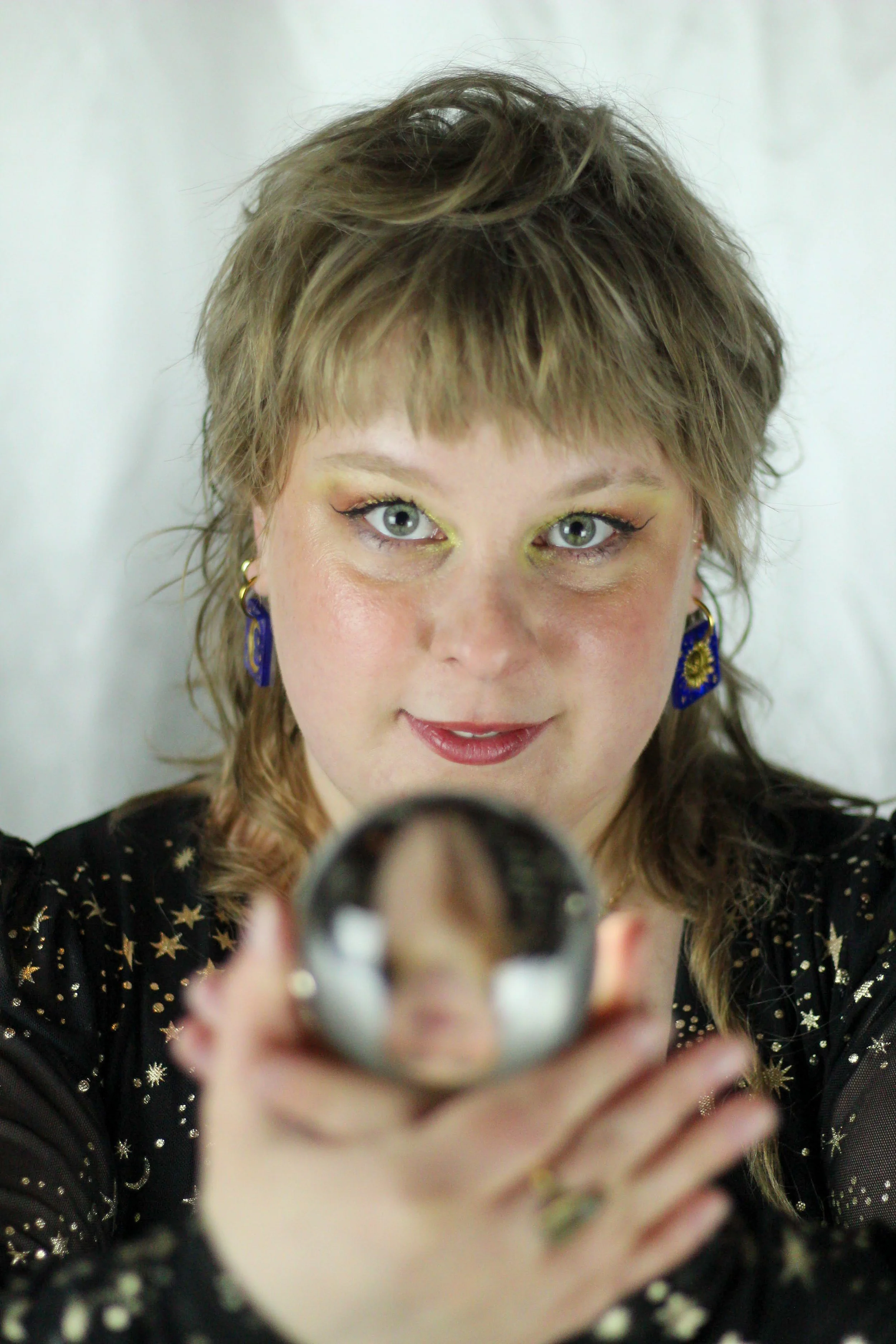 beautiful lesbian woman with blue eyes and whimsy goth aesthetic holding a crystal ball prism with  yellow golden eye makeup and a black dress with golden stars, looking at the camera in a playful whimsical way