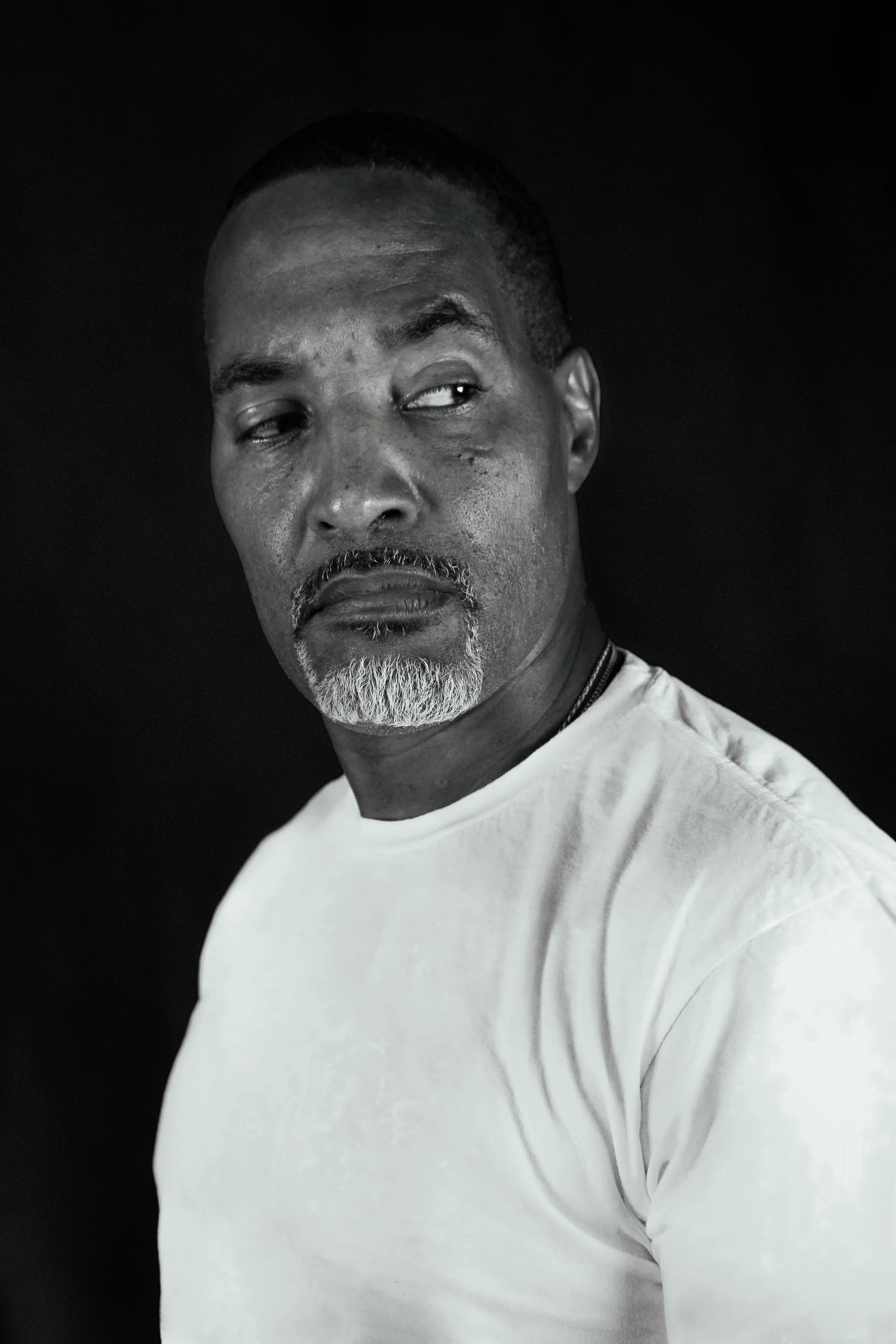 Black and white portrait of a middle-aged man with short hair and a beard, wearing a white t-shirt, looking to the side against a dark background. Looking like he is about to square up or squabble up with someone.