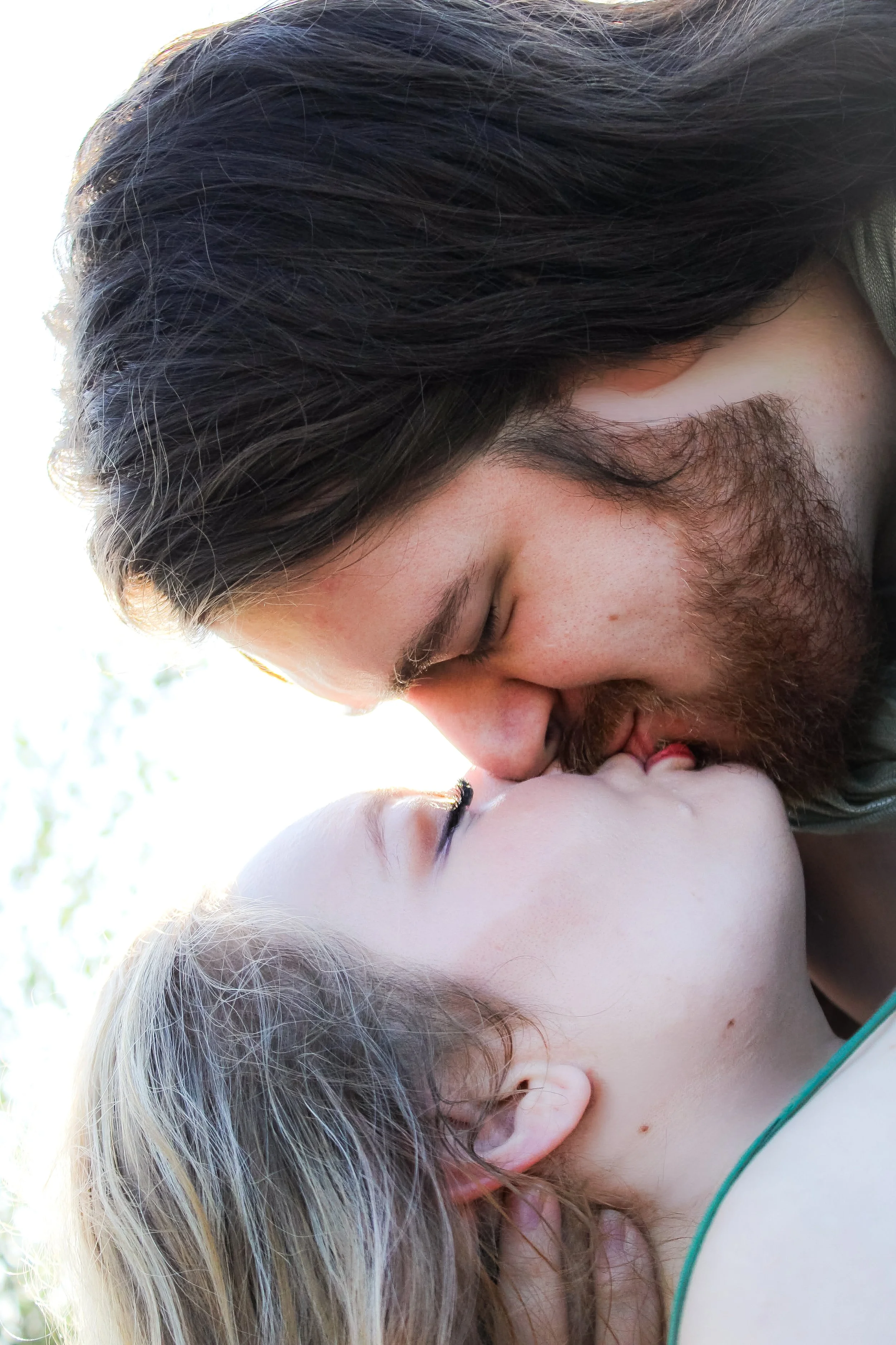 A man and woman sharing a kiss outdoors, with the man having long brown hair and a beard, and the woman with blonde hair.
