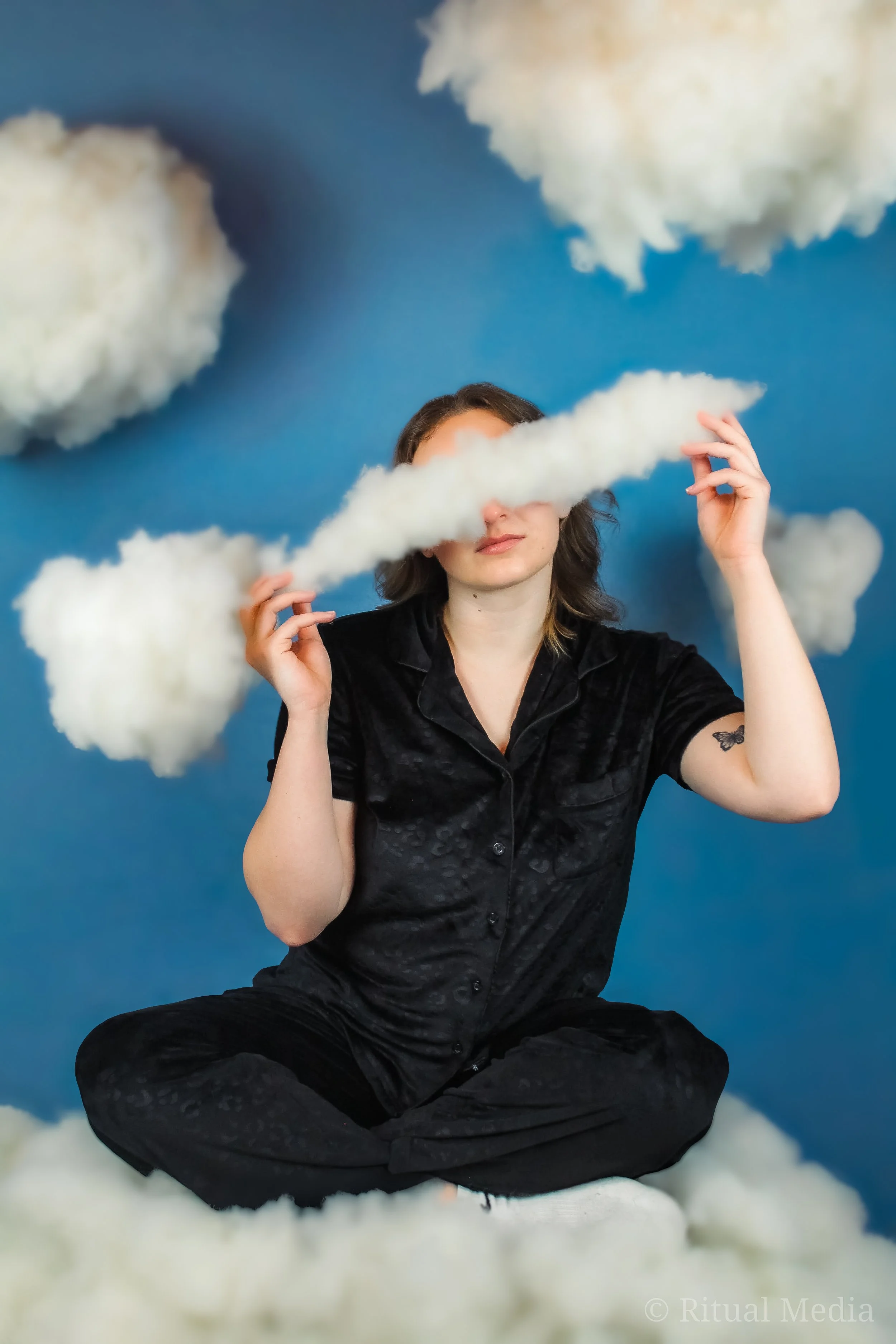 woman sitting on clouds with clouds covering her eyes giving a head in the clouds vibe, also a clouded vision idea here, suggesting the girl is daydreaming with her black silk pajamas and butterfly tattoo also has a whimsical and cartoonish vibe