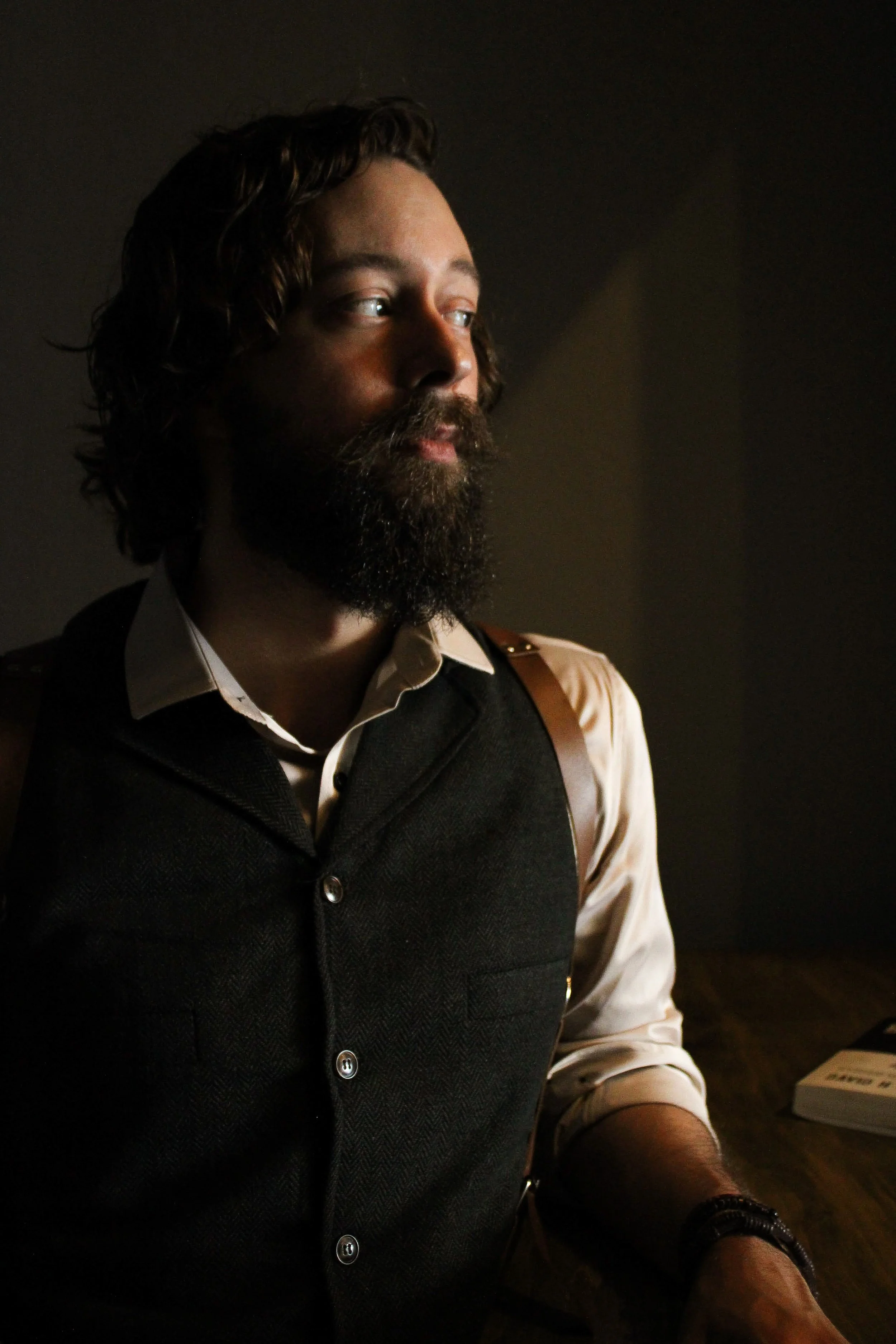 A man with a beard and long hair sitting at a table, looking thoughtful in low lighting.