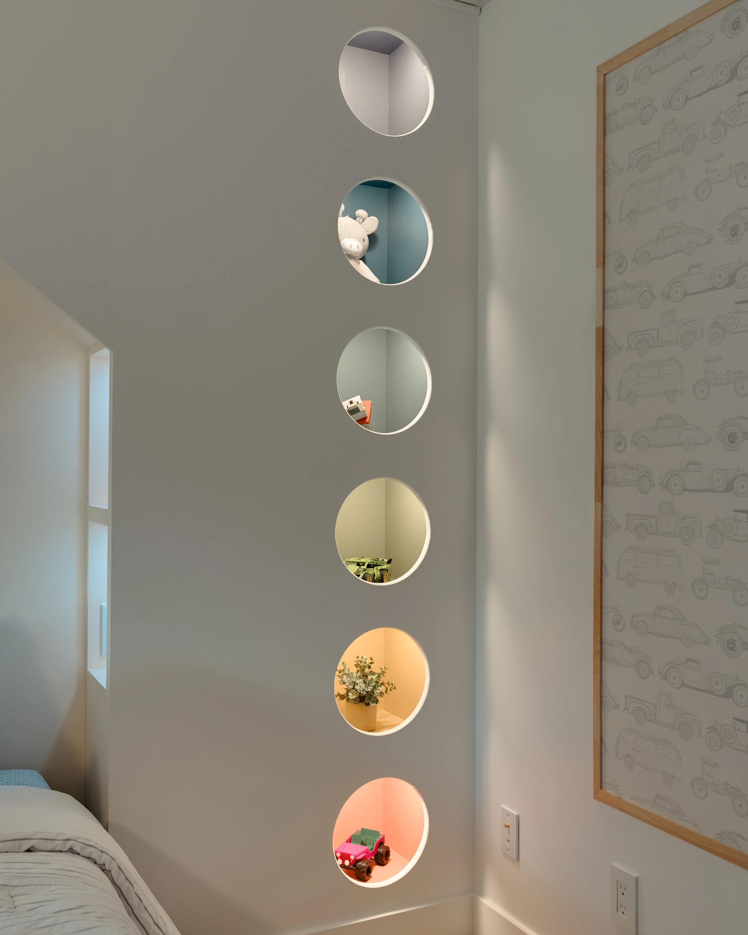 Mur avec cinq trous ronds percés, chaque trou contient un objet décoratif ou une lampe de couleurs différentes, dans une chambre. Sur la droite, un mur avec un motif de voitures, prise électrique en bas.