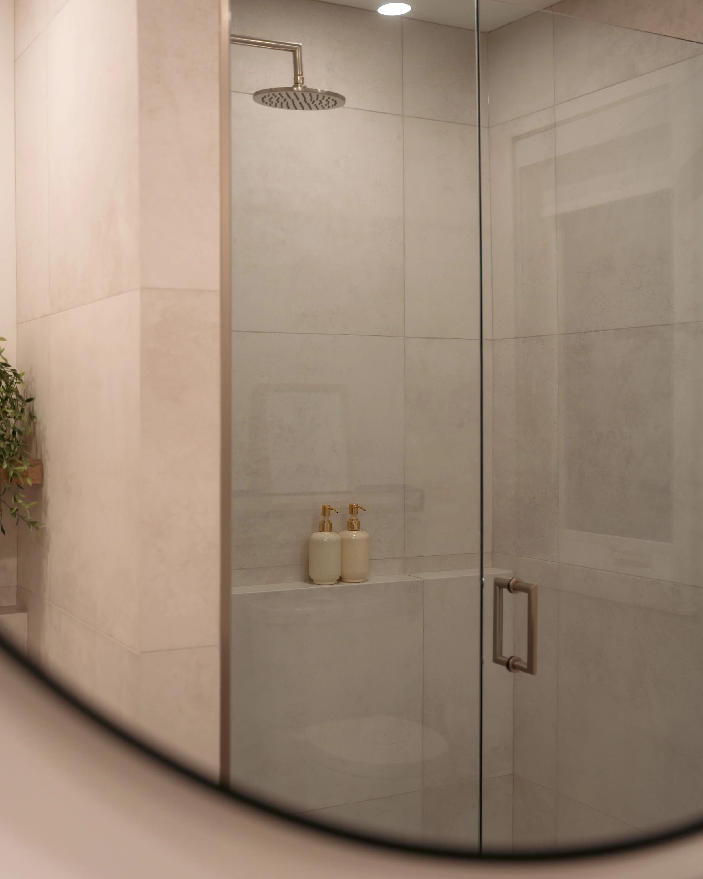 Douche à l'italienne avec porte en verre, parois en carrelage beige clair, deux distributeurs de savon ou shampoing, éclairage au plafond, miroir visible à droite, ambiance moderne et épurée.