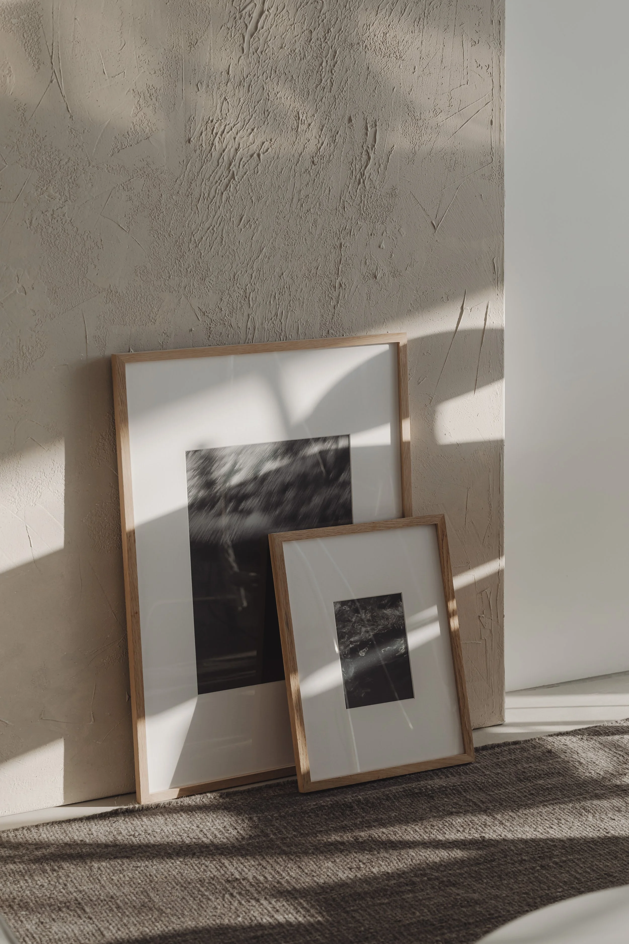 Deux cadres photo noirs et blancs avec des bords en bois, posés contre un mur en plâtre texturé, avec des rayons de lumière traverse de petites fenêtres.
