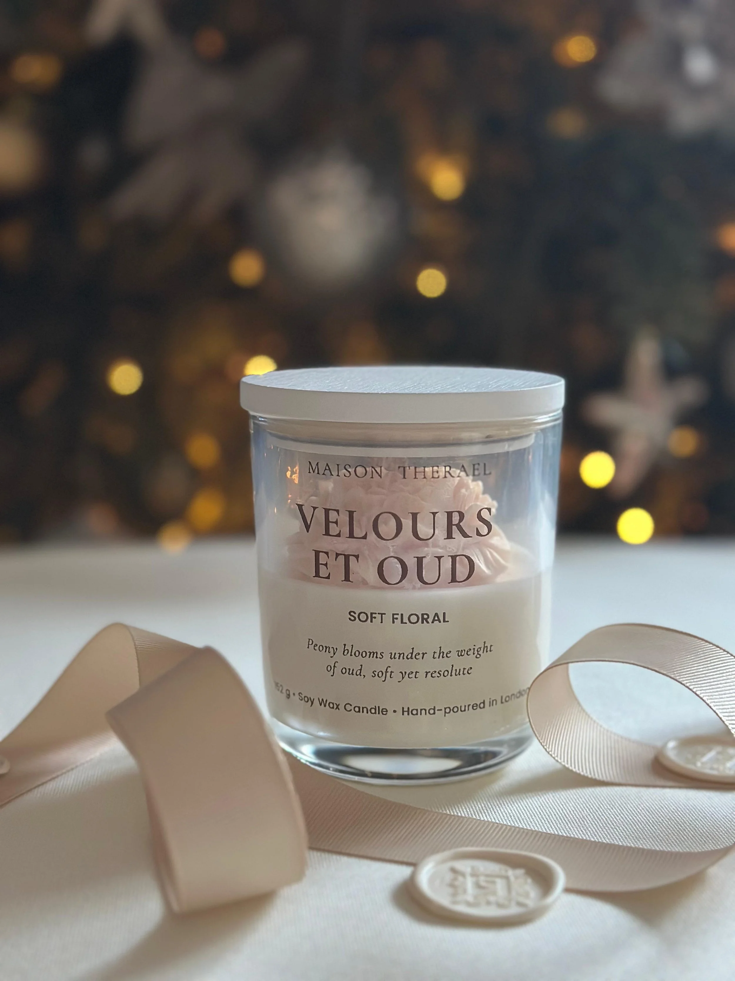 A glass jar candle with a white lid, labeled 'Velours et Oud' by Maison Therael, placed on a white surface with a cream-colored ribbon and wax seal, with a blurred Christmas tree in the background.
