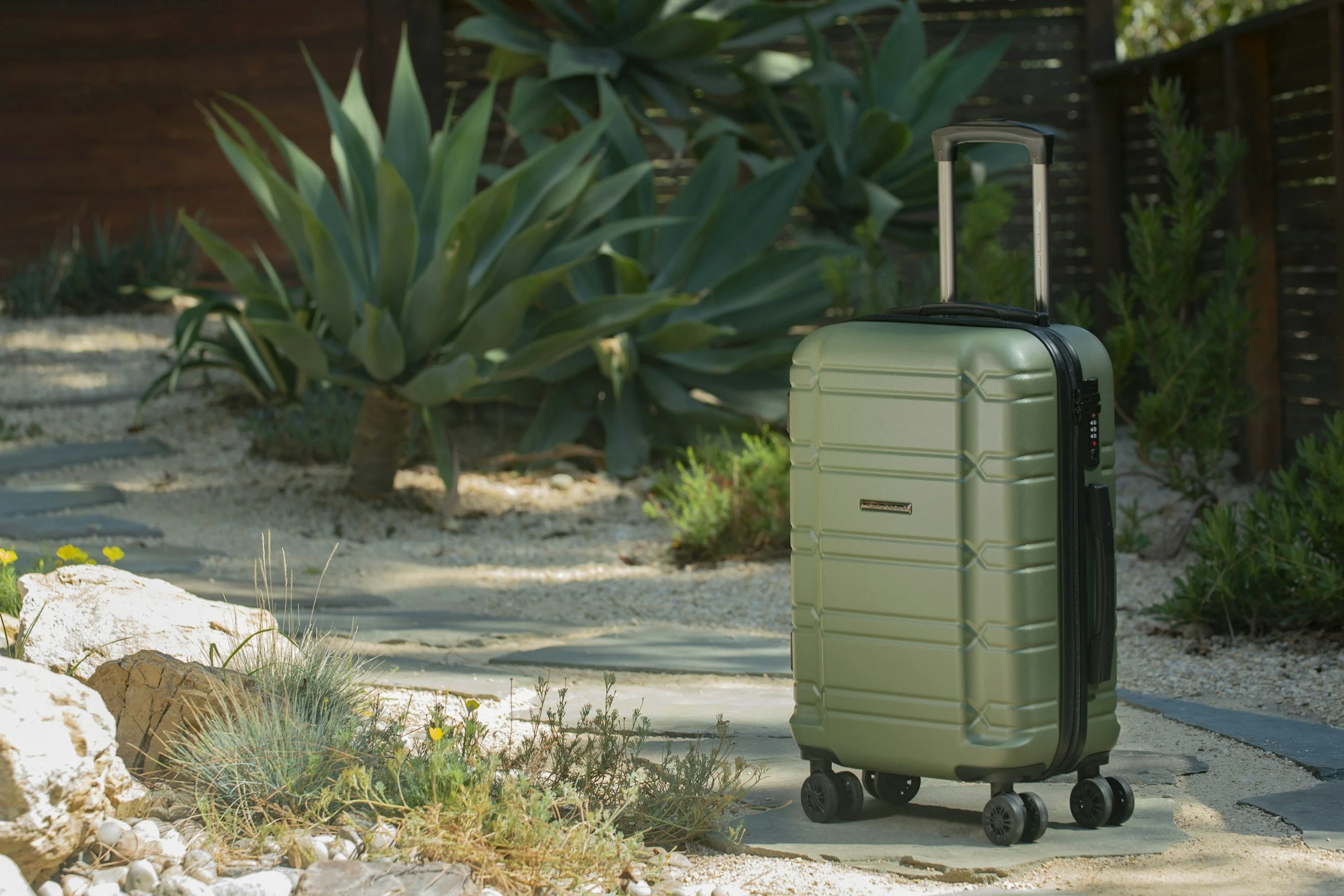 A green hard-shell suitcase with four black wheels and an extended handle standing on a stone pathway in a backyard with large agave plants, small yellow flowers, rocks, and a wooden fence in the background.