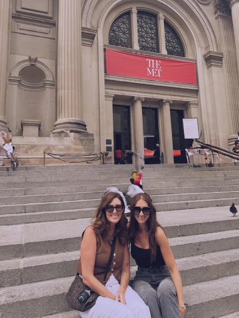 Two women sitting on stairs outside a building with large columns, entrance, and a red sign that reads 'The Met'.