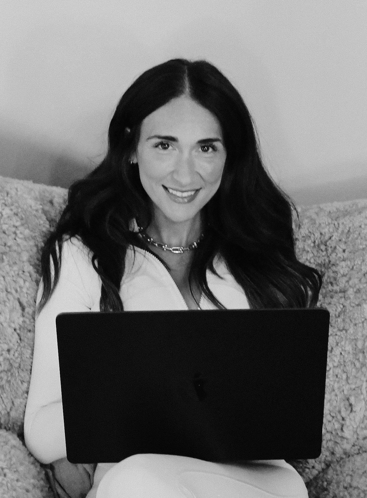 Black and white photo of a woman with long dark hair smiling while sitting on a couch with a laptop on her lap.