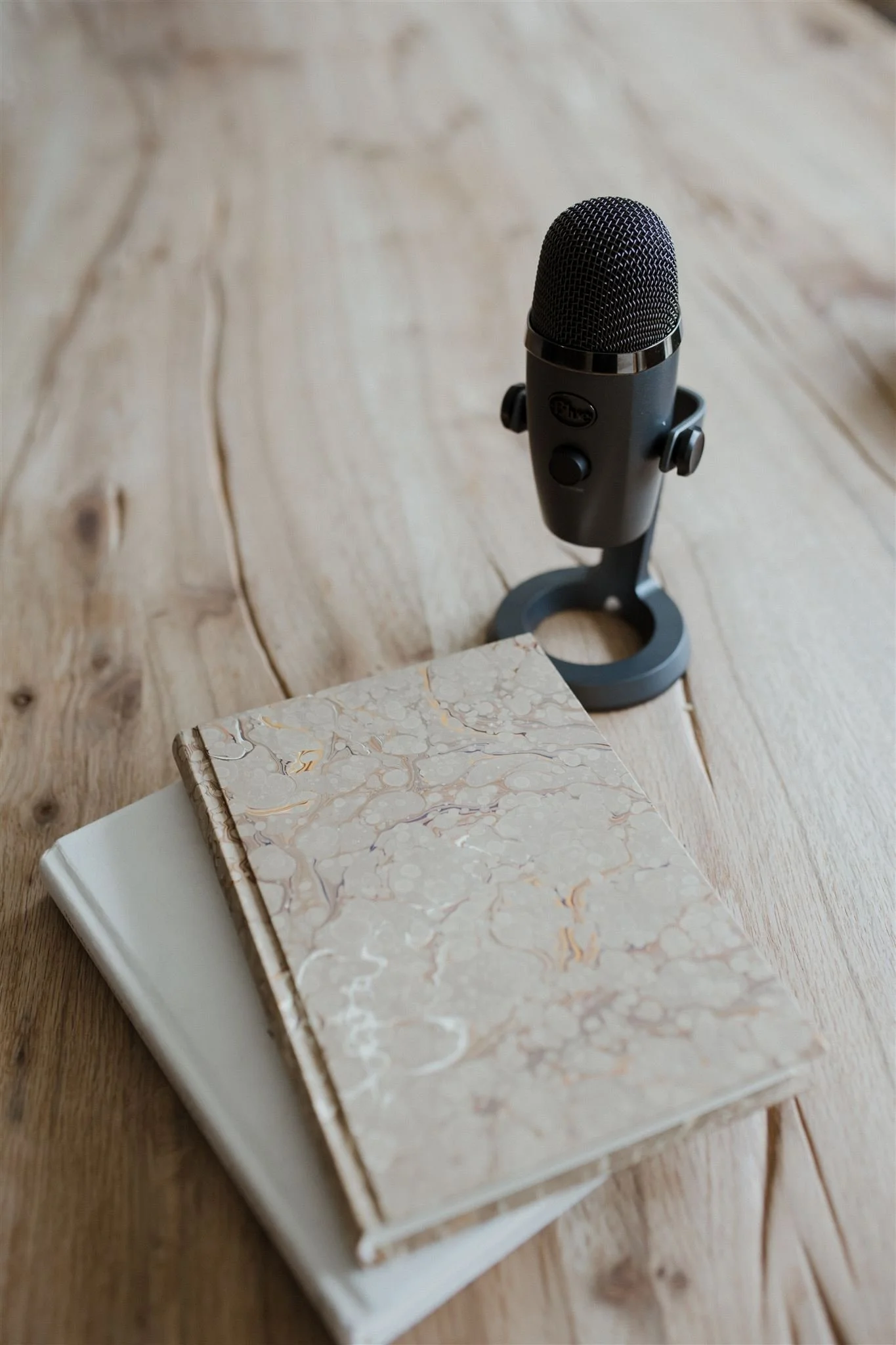 A black microphone on a wooden surface next to two books, one with a textured beige cover and the other with a plain white cover.