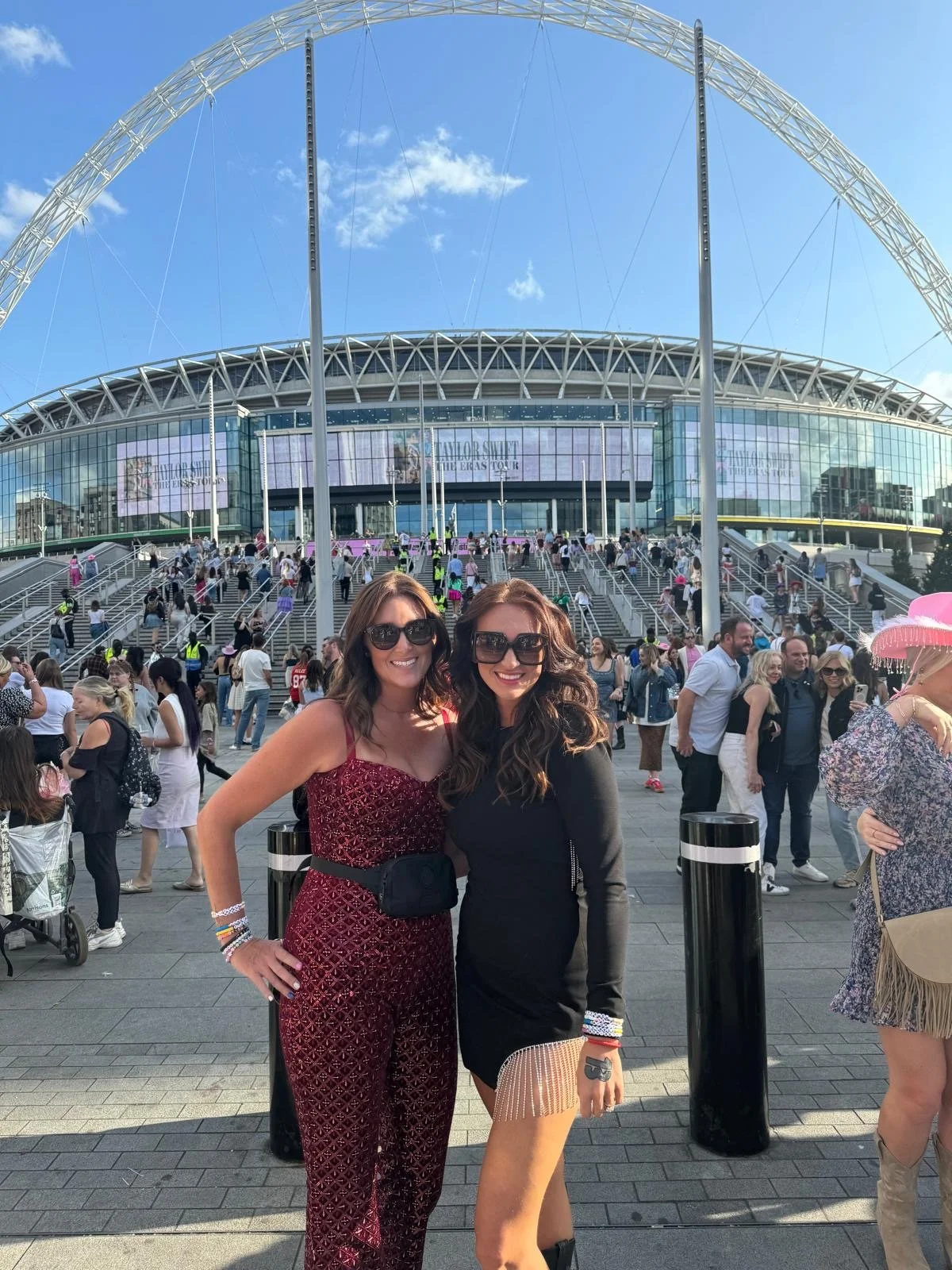 Two women smiling and posing in front of Tottenham Hotspur Stadium during a sunny day with many people in the background.