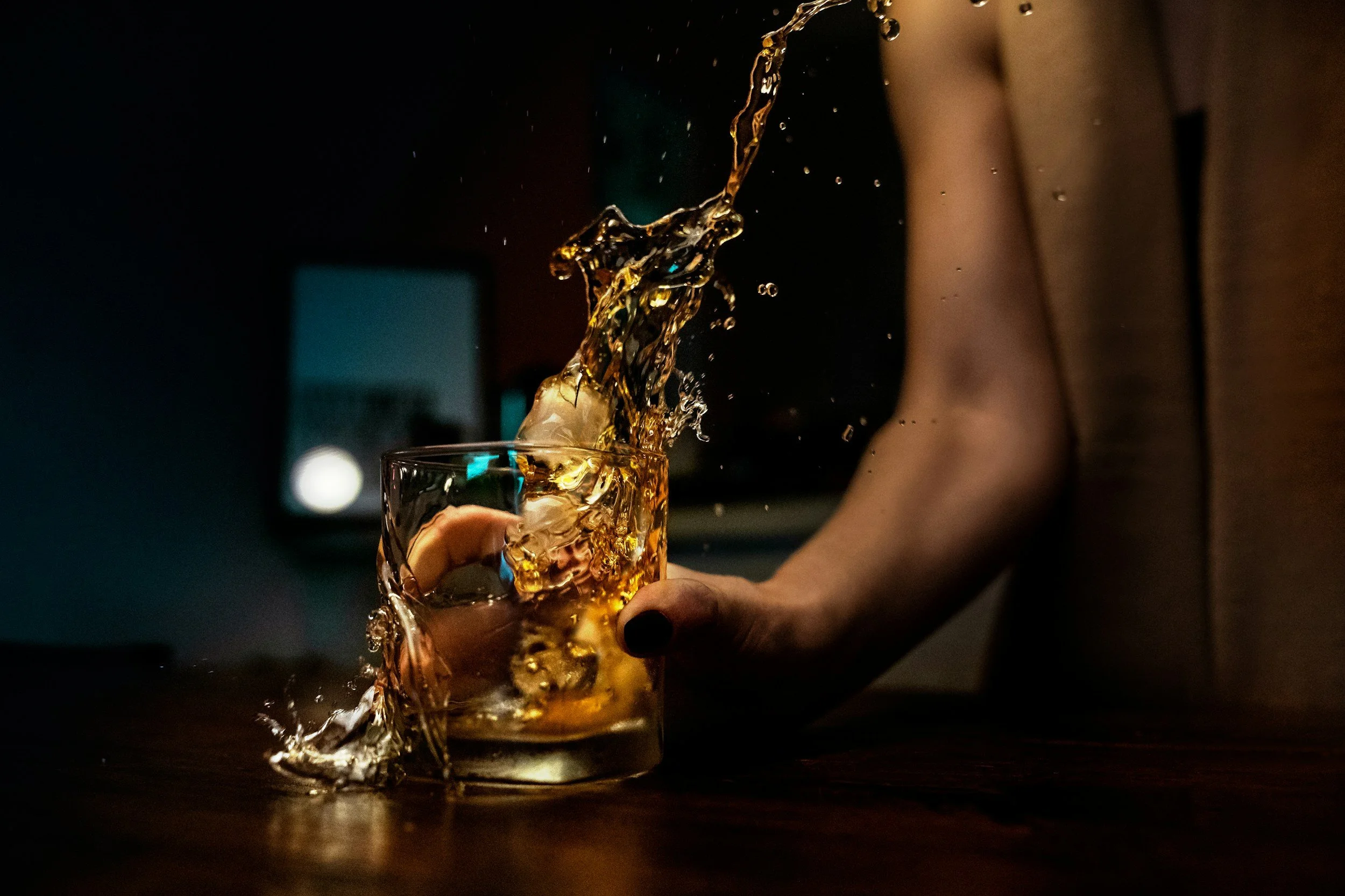 Person holding a glass of whiskey as liquid spills out in a dark room.