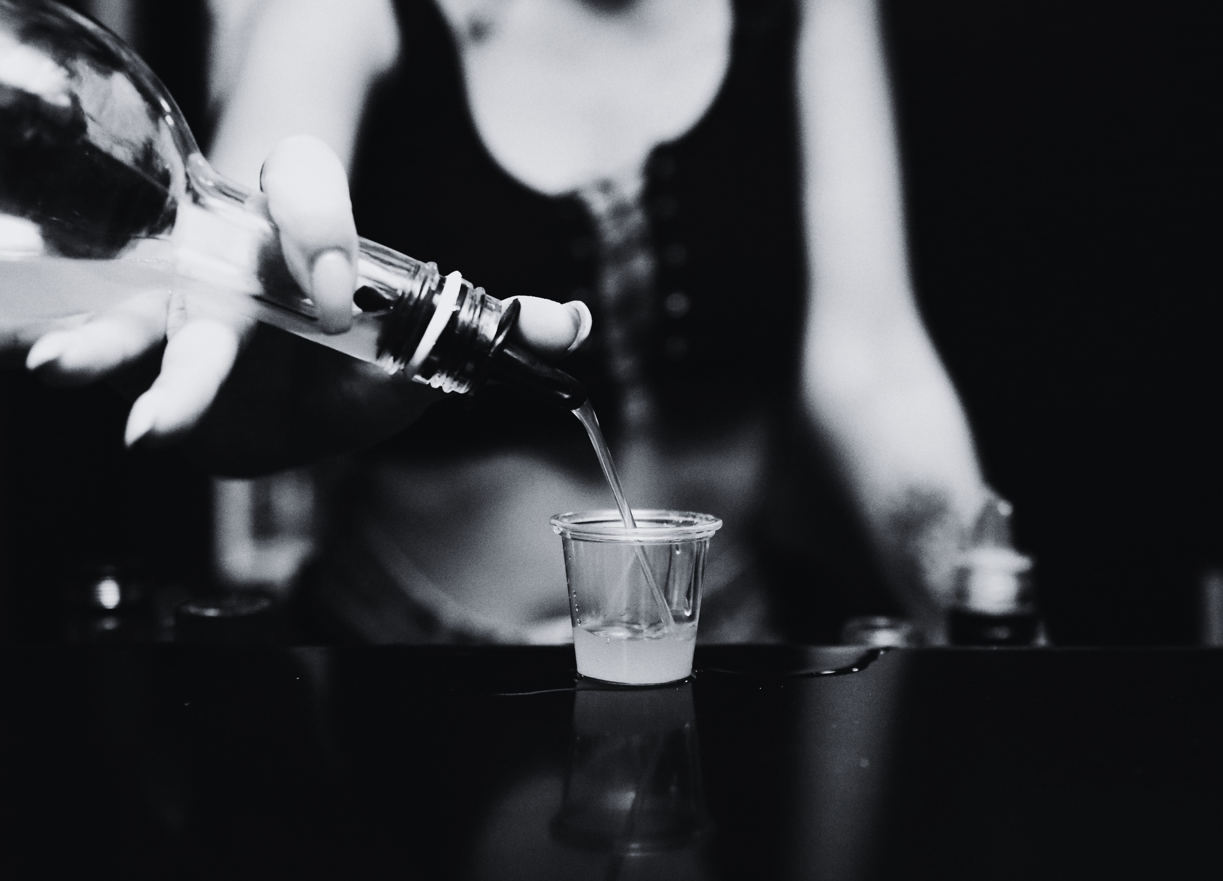 A person pours a liquid from a glass bottle into a small clear plastic cup on a dark surface, with a blurred background.