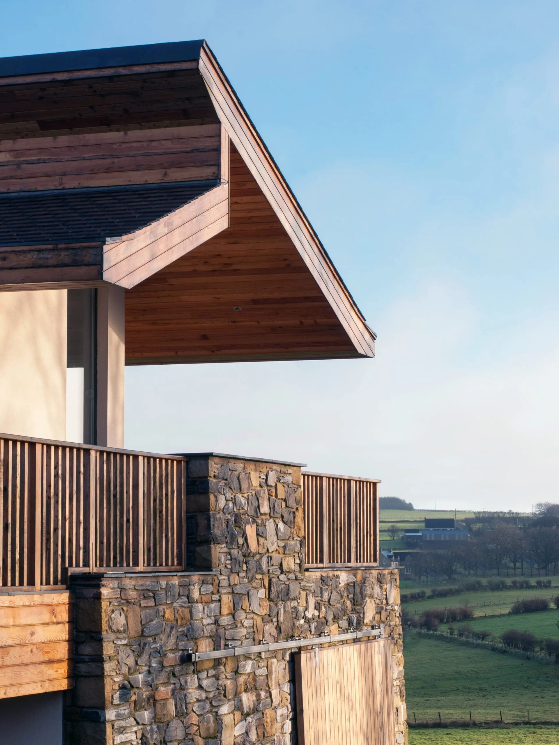 Close-up view of a modern house with a stone wall, wooden balcony railing, and an overhanging wooden roof against a blue sky and distant green landscape.