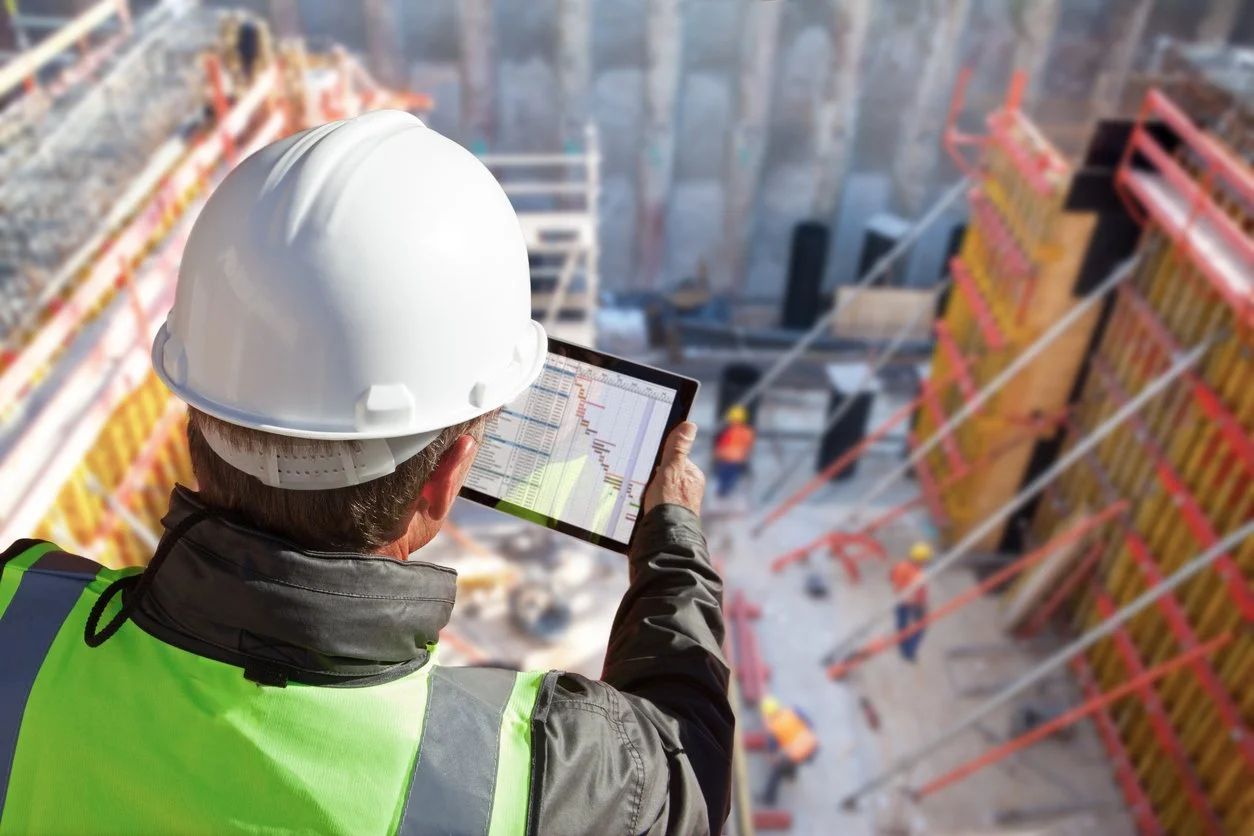 Construction worker in safety helmet and vest looking at a tablet with construction site plans or charts, background shows a multi-story building under construction with scaffolding and workers.
