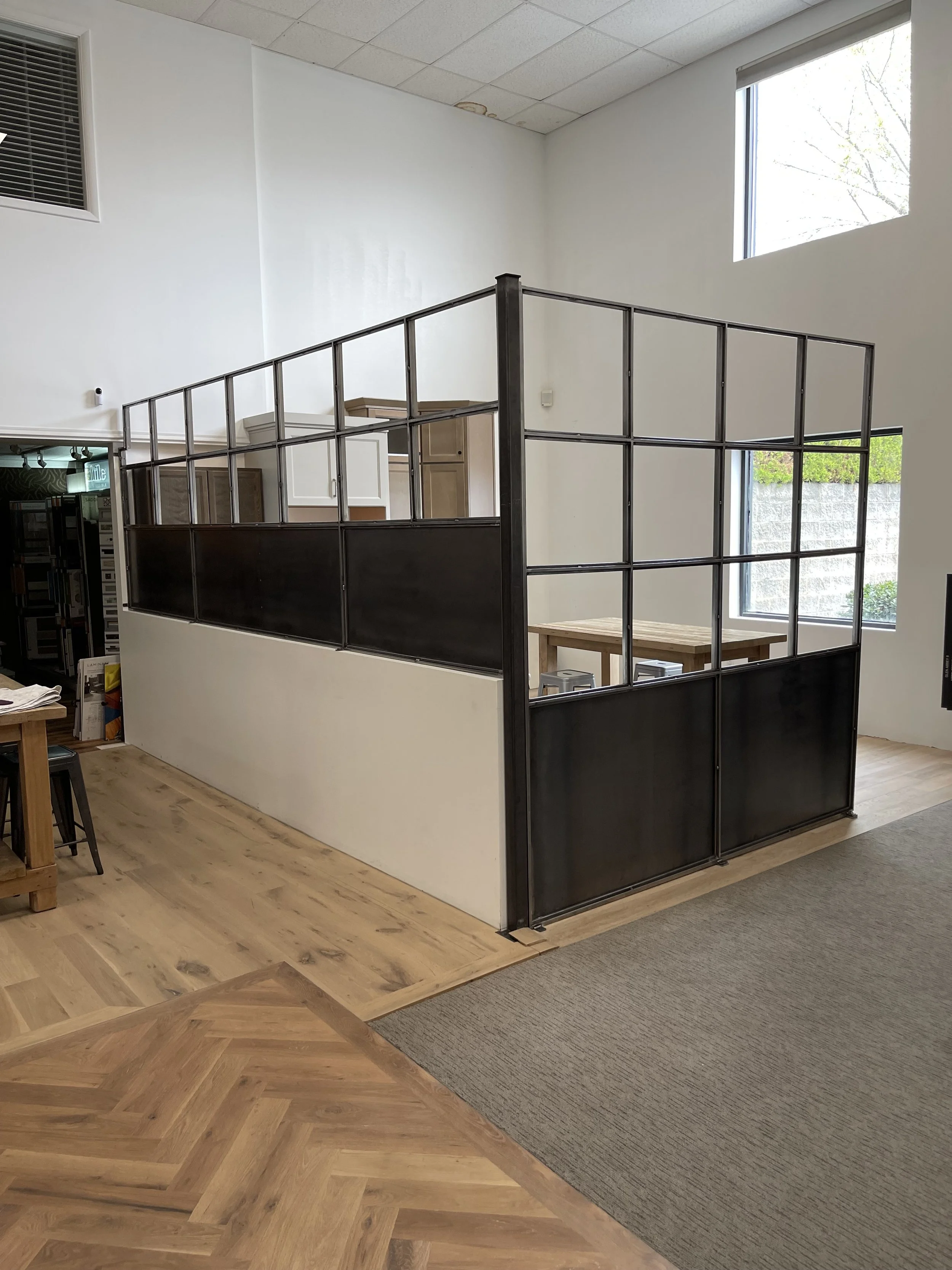 Indoor space with a loft or mezzanine level enclosed by a black metal frame and panels, with wooden flooring and large windows letting in natural light.