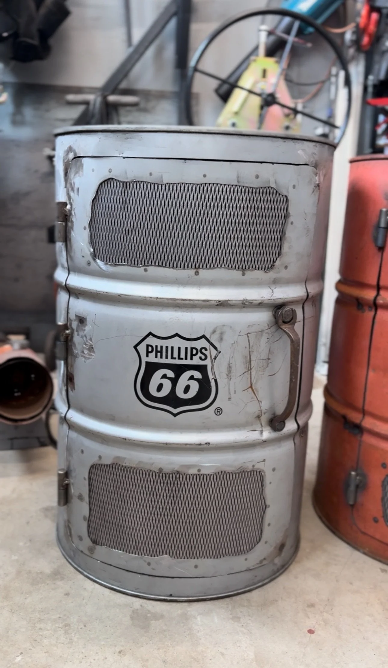 Vintage metal trash can with a Phillips 66 shield logo, two metal mesh panels, and rustic paint, placed on a concrete floor in a garage or workshop.