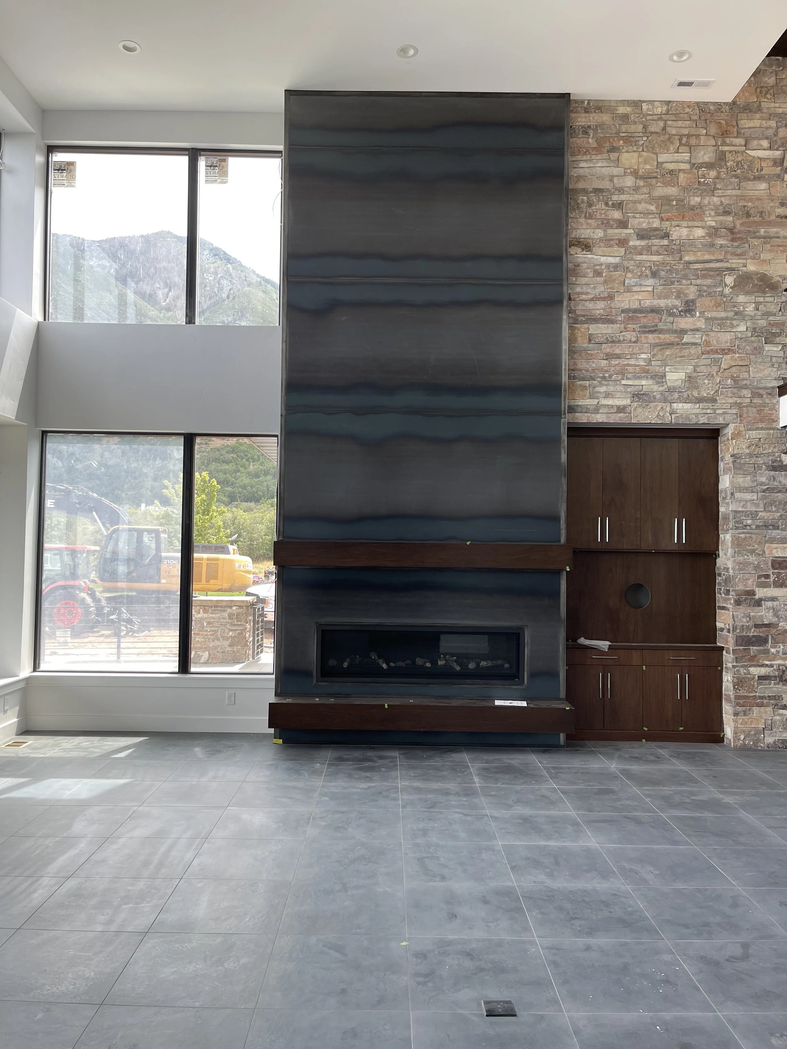 Interior of a modern living room with a tall fireplace, brick wall, large windows showing a mountain view, and built-in wooden cabinets.