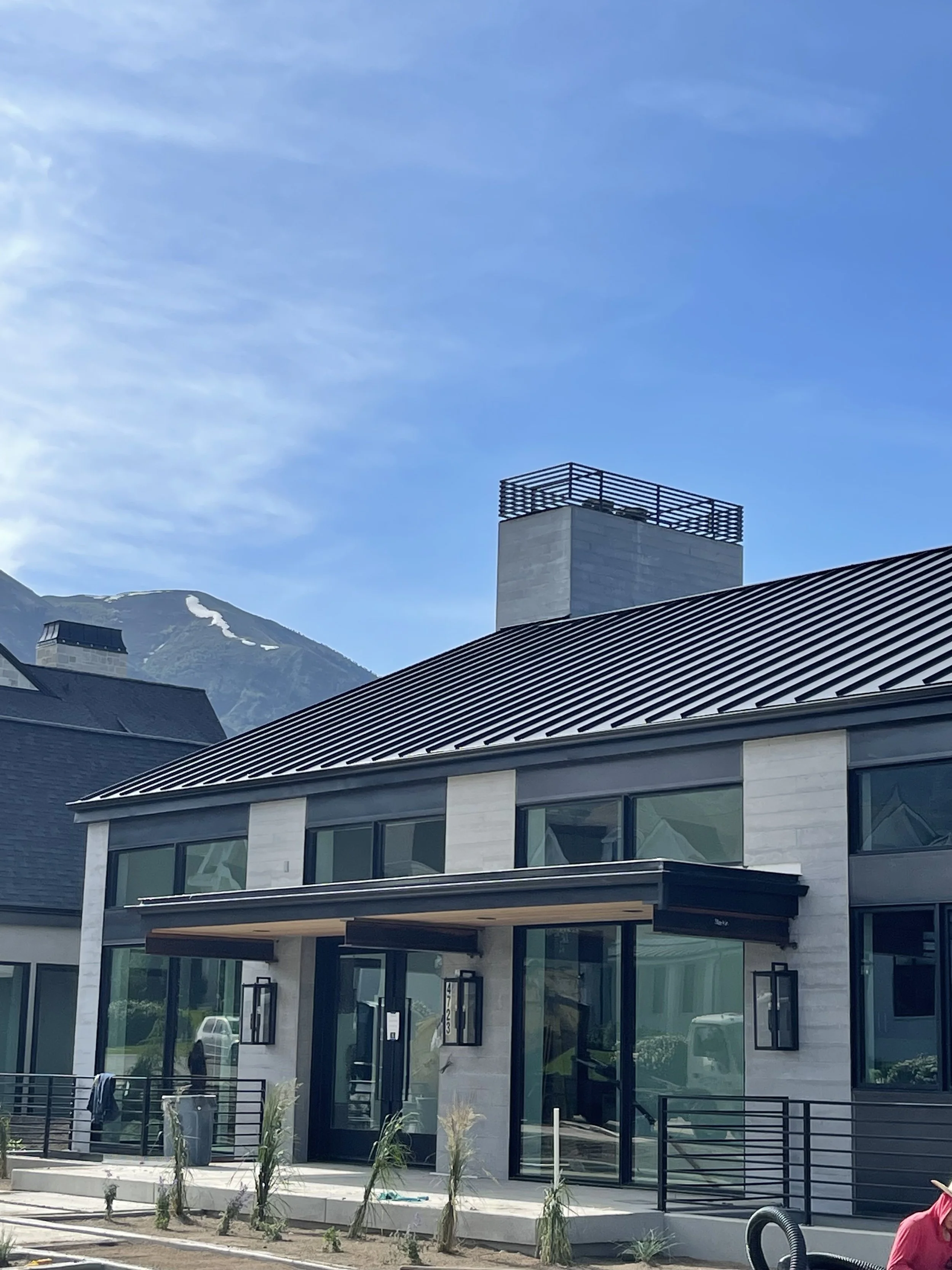 Modern residential house with large glass windows, a metal roof, and a balcony with a railing, set against a mountain backdrop with a blue sky.