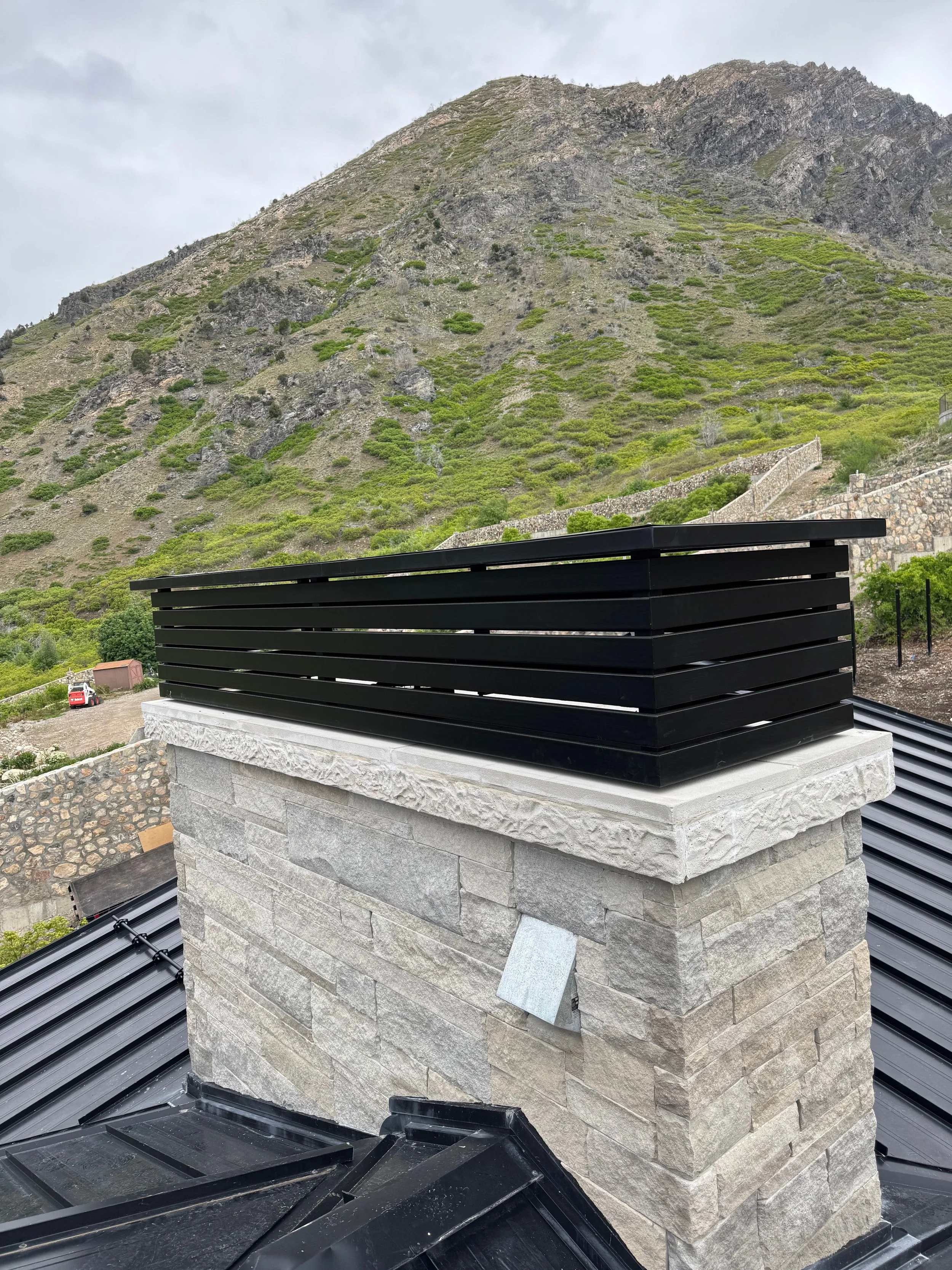 Close-up of a stone chimney with a black metal cap on a roof, with a mountainous landscape in the background.
