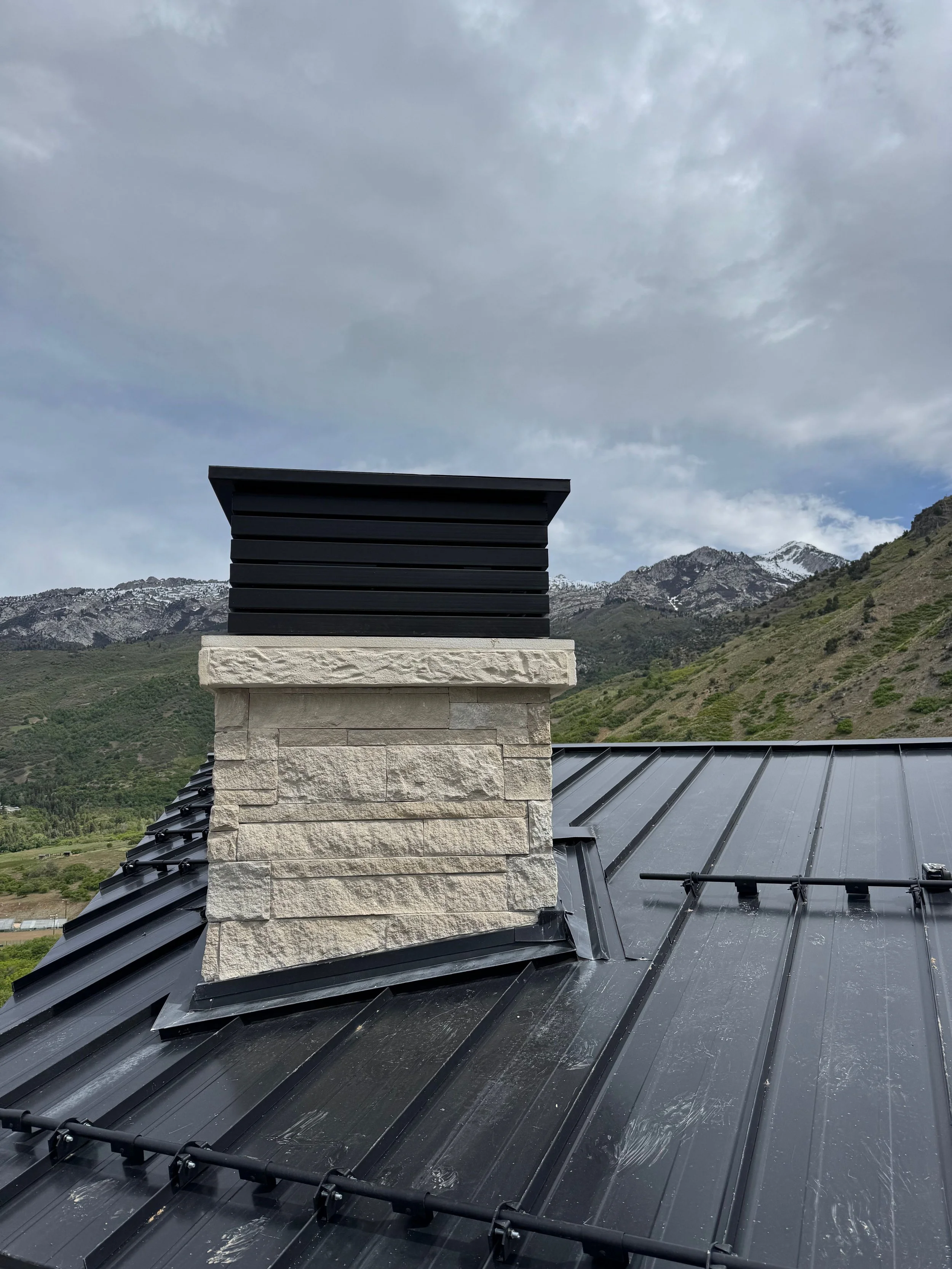 Close-up of a chimney with a black metal cap on a rooftop with black metal roofing, mountains, and a cloudy sky in the background.