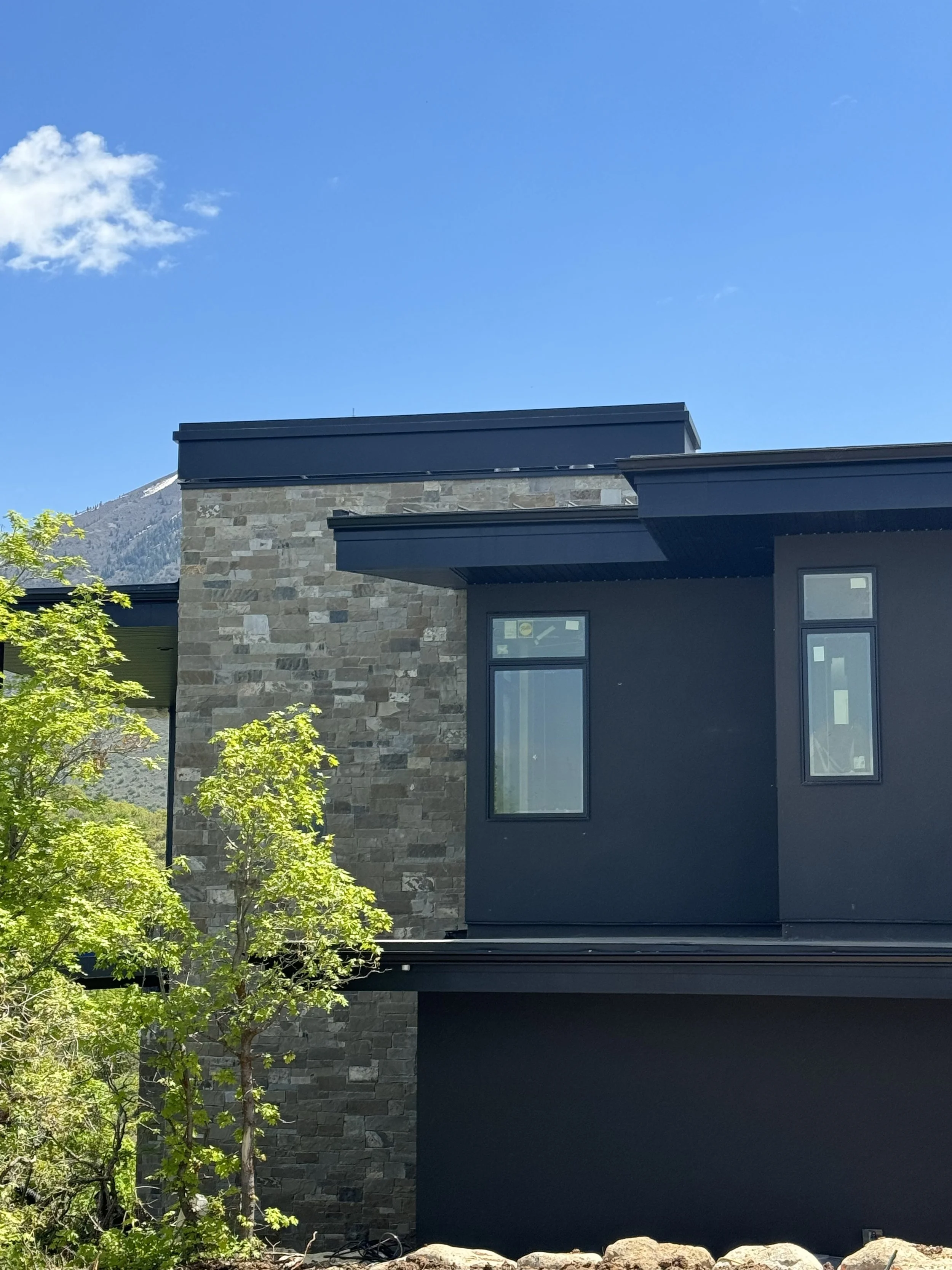 Part of a modern house with stone and black siding, multiple windows, and a sloped roof, with green trees and mountains in the background under a blue sky.