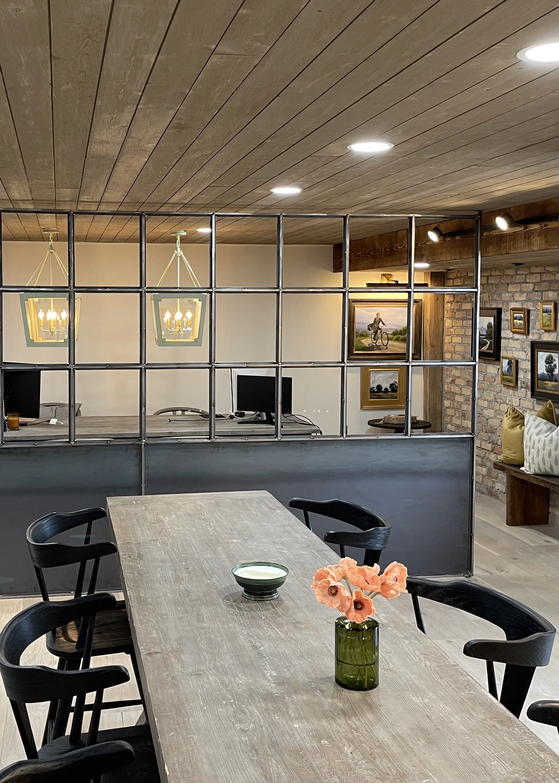 A dining room with a wooden table and black chairs, a vase with pink flowers, and a wall with framed pictures. Behind a metal divider, an office area with computers and wall art is visible. The ceiling has wooden panels and recessed lighting.