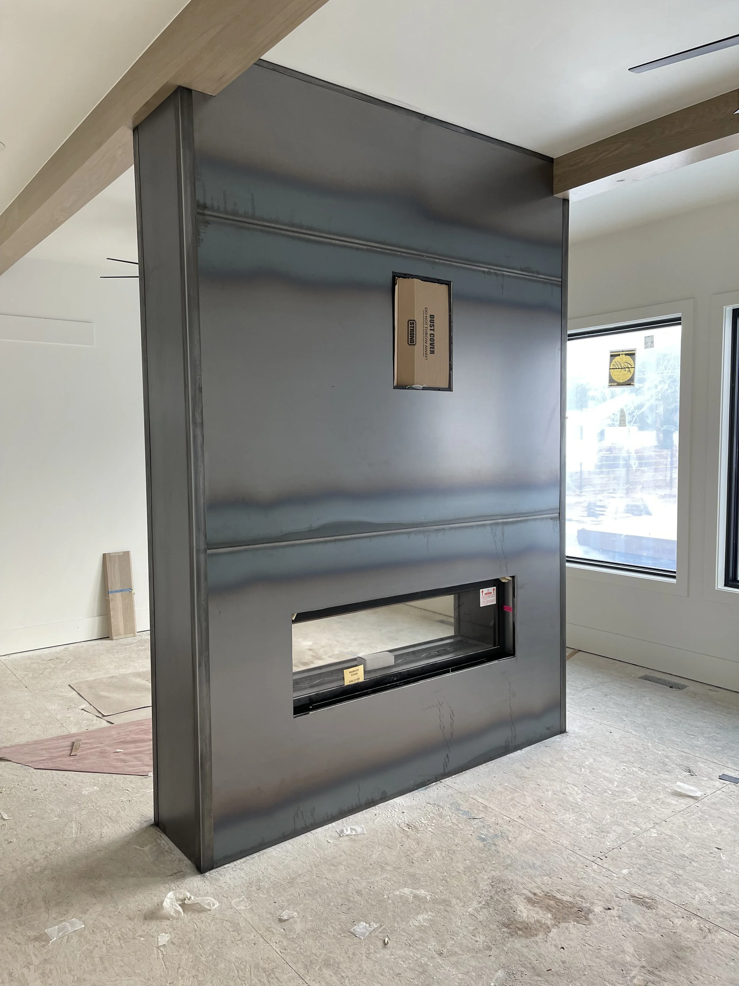 A partially installed modern fireplace with a box-like metal frame and a rectangular glass viewing window, situated in a room under construction.