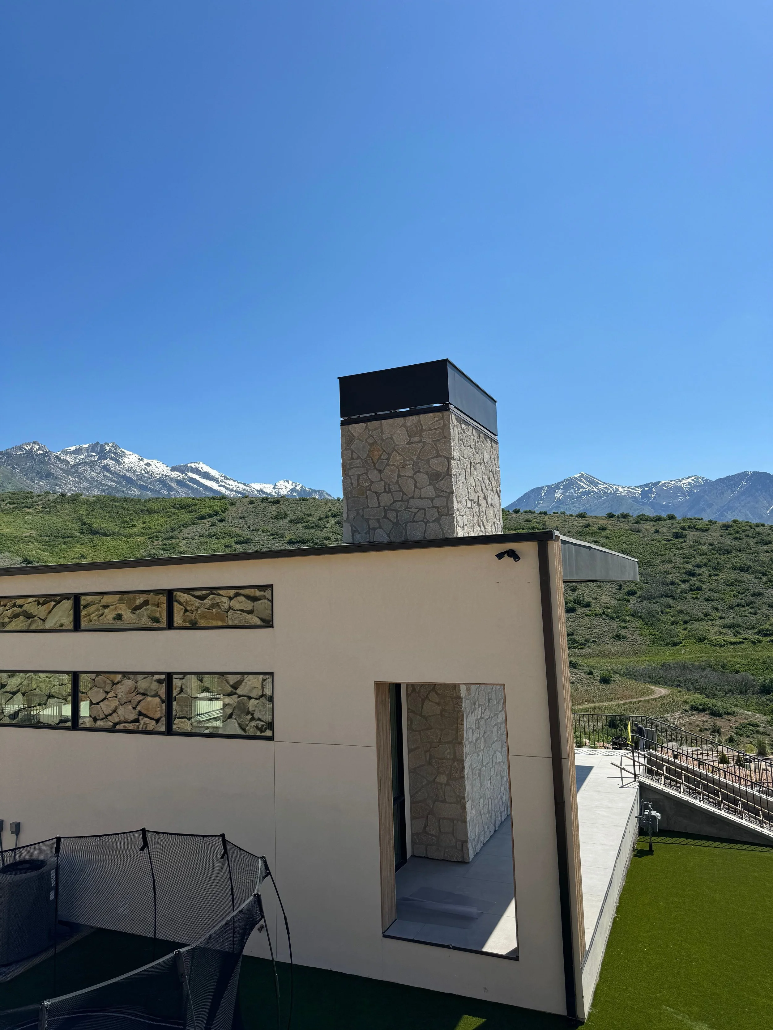 Modern house with stone accents, large windows, and a chimney, set in a mountainous landscape with snow-capped peaks in the background under a clear blue sky.