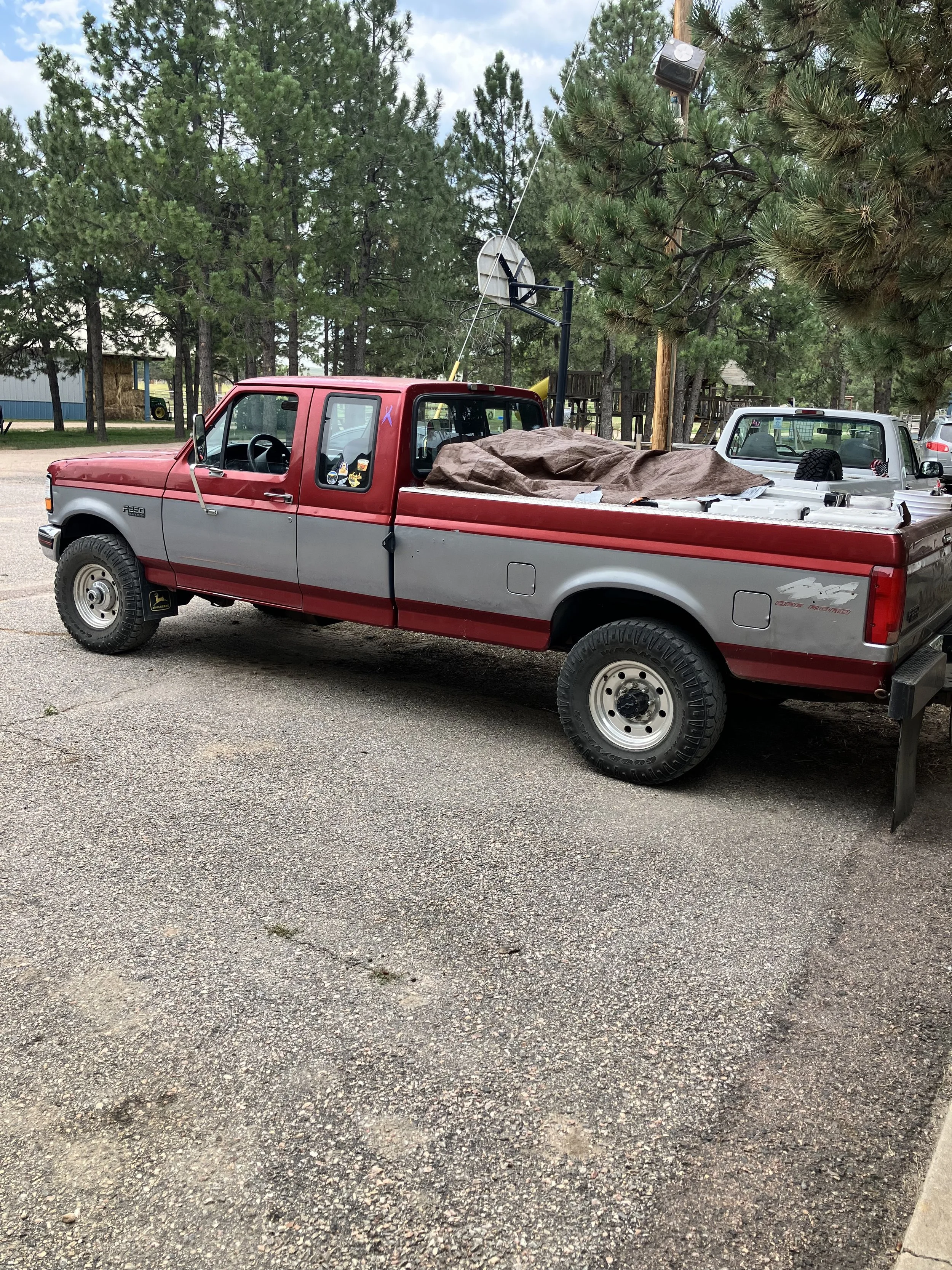 Red and gray pickup truck parked on a gravel surface with trees and a basketball hoop in the background.