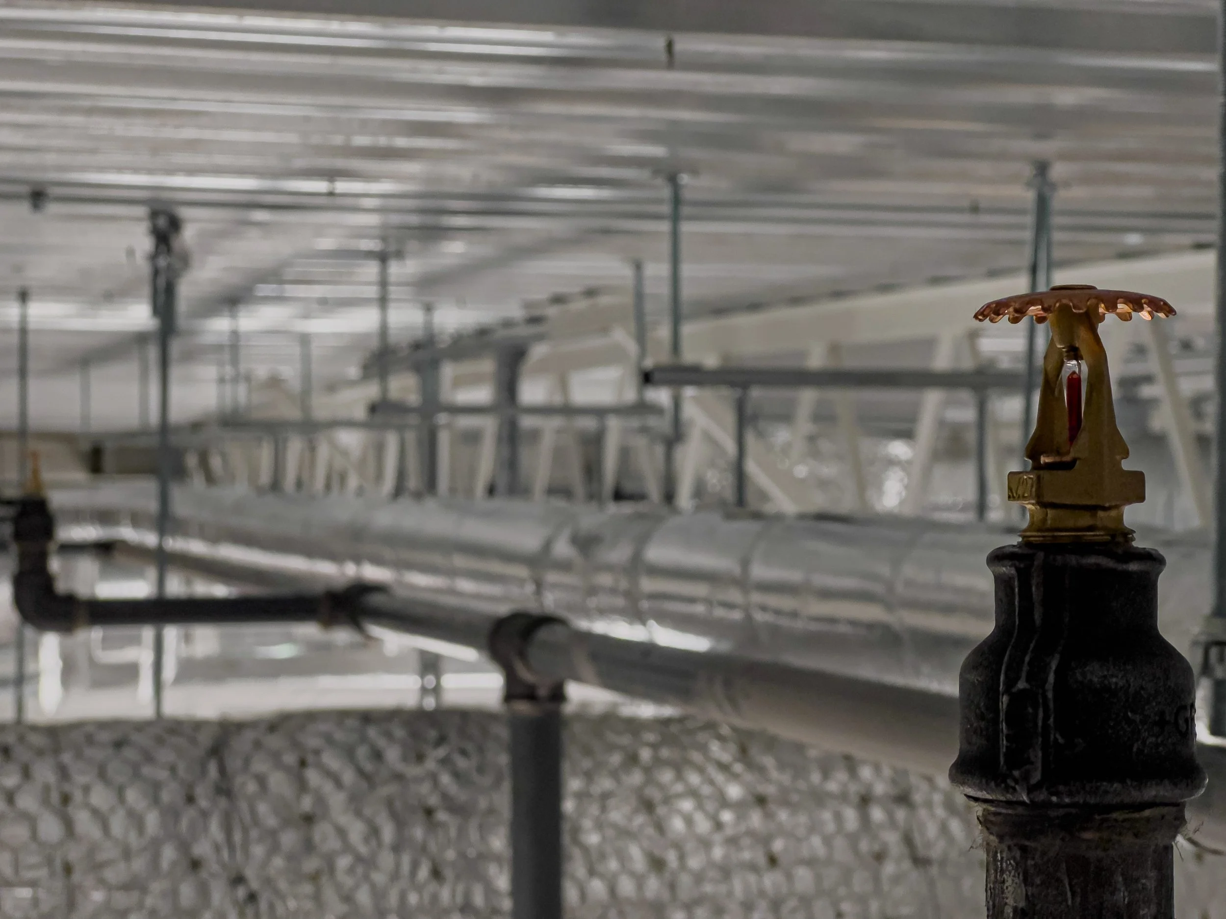 Close-up of a fire hydrant with a fire department connection, situated in an industrial setting with pipes and metal scaffolding in the background.