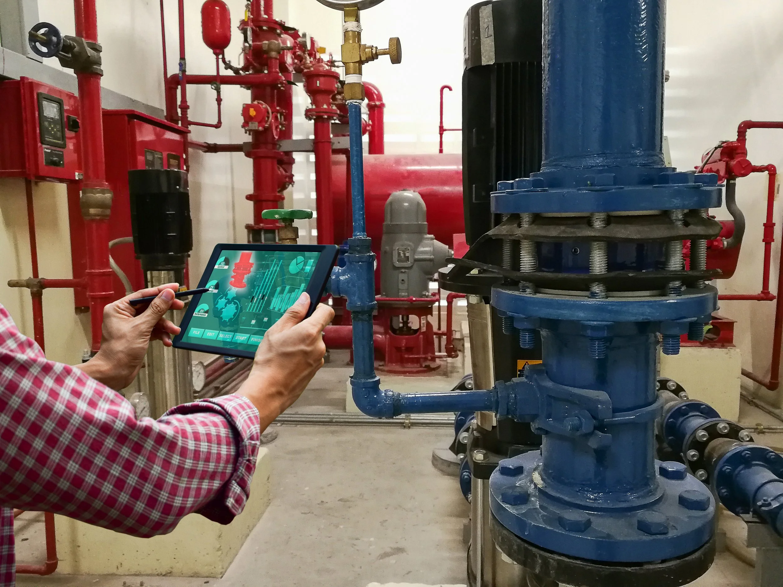 Person in a red checkered shirt holding a tablet with a digital interface, inspecting industrial pipes and machinery with red and blue pipes in an industrial or facility setting.