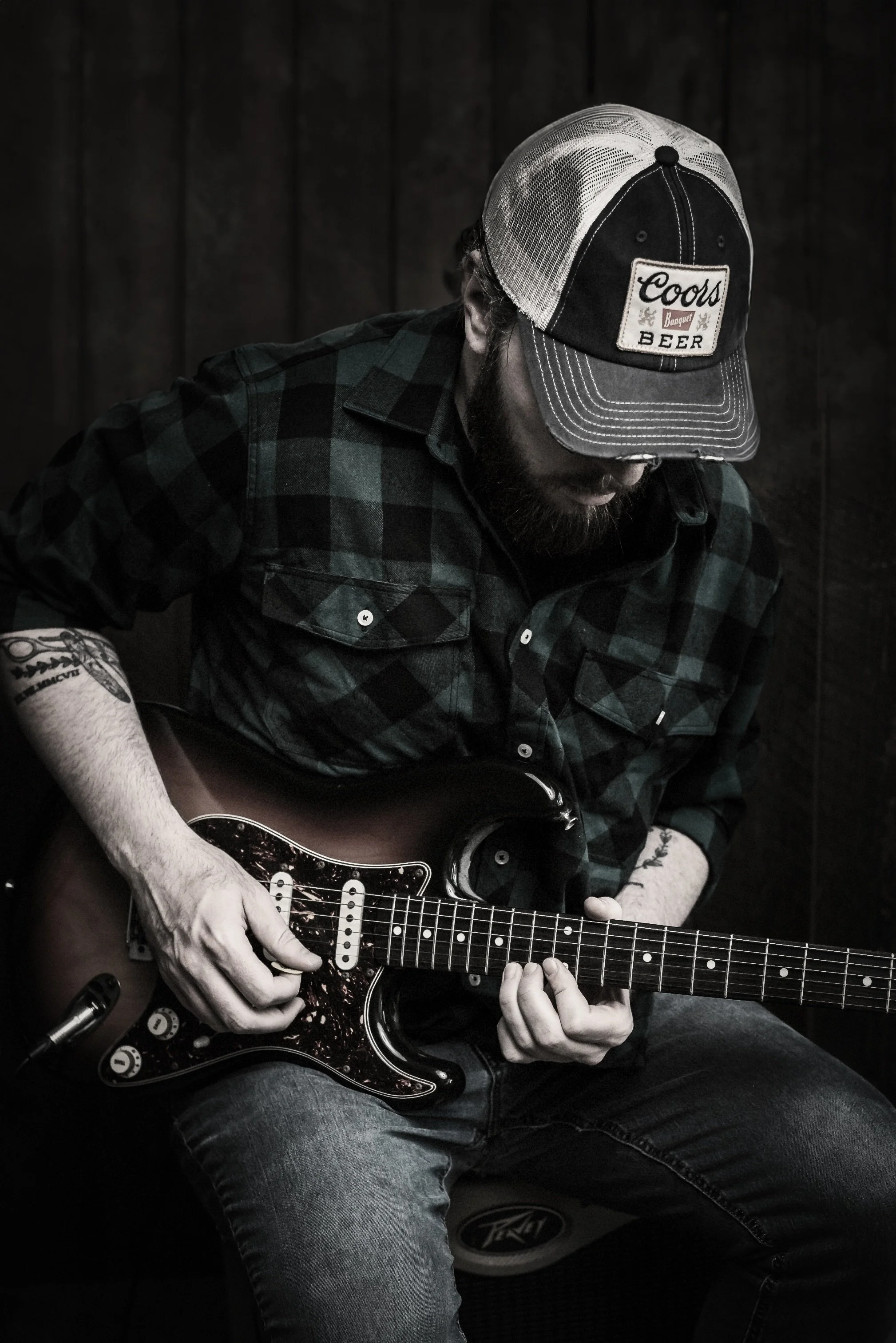 A man with a beard wearing a black and green plaid shirt, a trucker cap labeled 'Coors Beer,' playing an electric guitar, sitting against a dark wooden background.