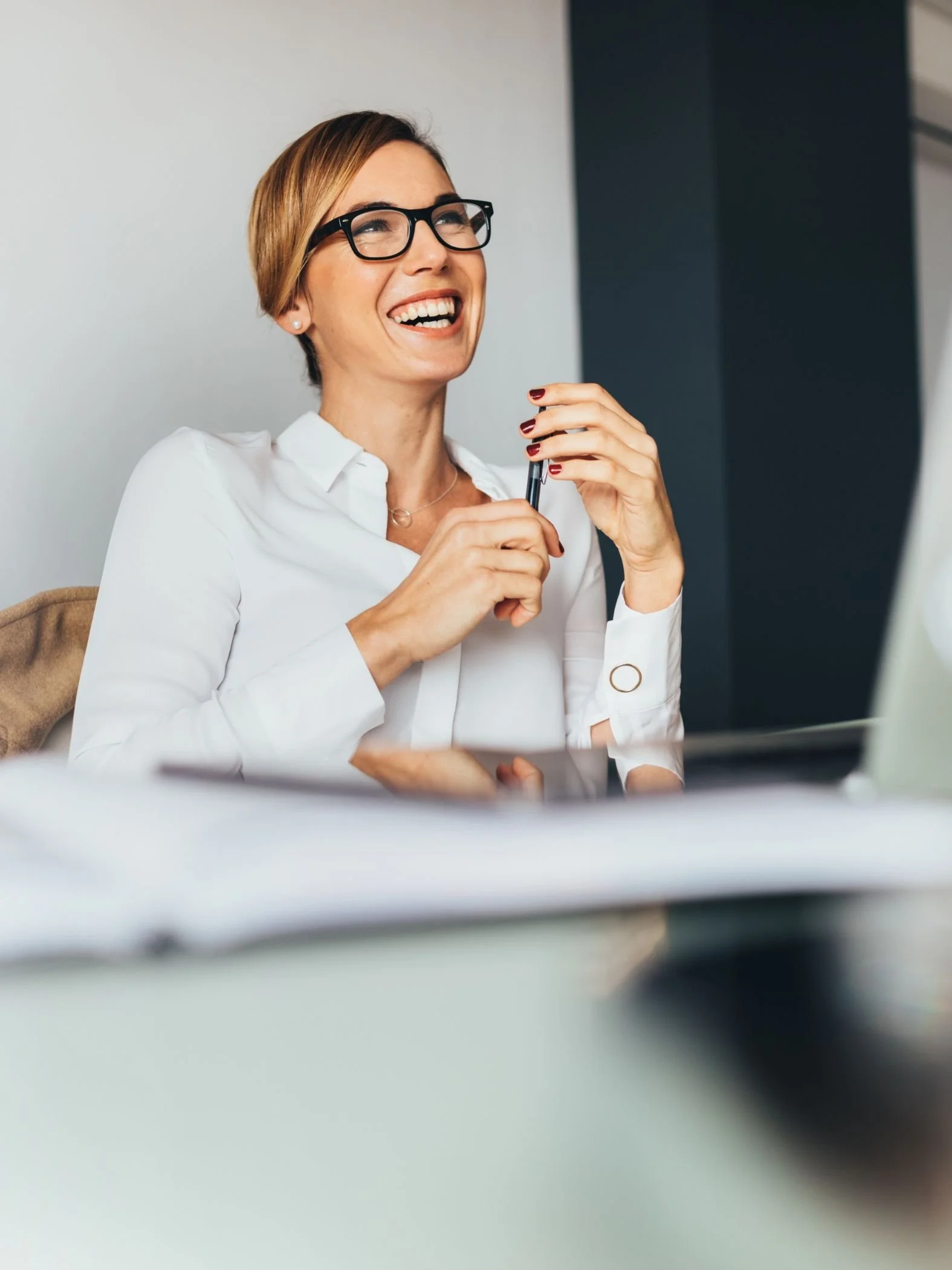 A woman with short hair, glasses, and a white shirt smiling and holding a pen, sitting at a desk.